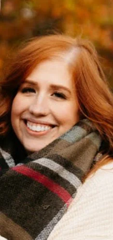 A smiling woman with red hair outdoors during fall, wearing a striped scarf.
