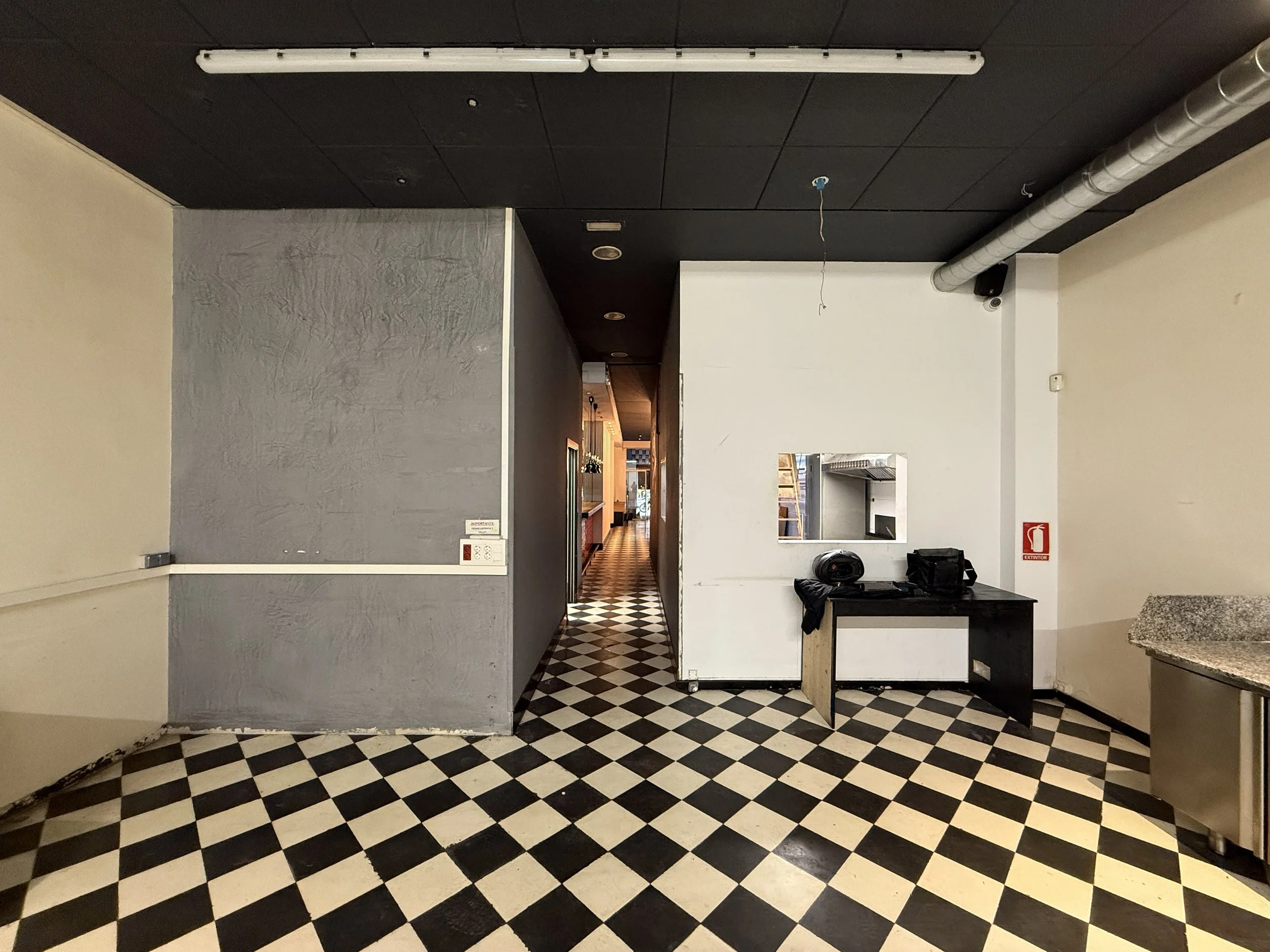 Interior de restaurante o cafetería con paredes blancas y grises, piso de baldosas en patrón de damero en blanco y negro, ampolletas colgando del techo, y una mesa negra con objetos encima, en un espacio aparentemente en reparación o cerrado.