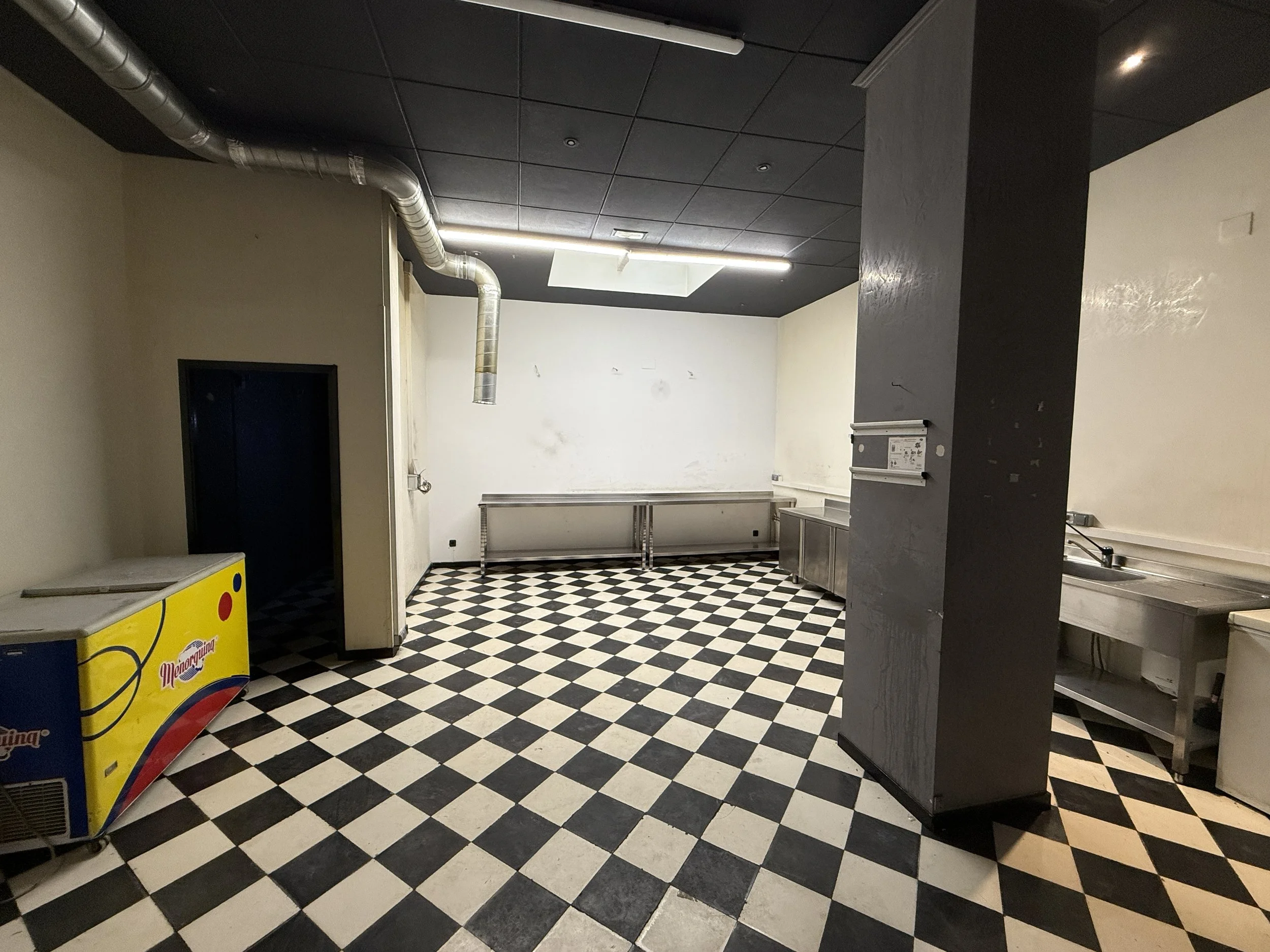 Cocina industrial vacía con baldosas de suelo en patrón de checkered blanco y negro, un refrigerador de hielo amarillo a la izquierda, mesadas de trabajo de acero inoxidable en el fondo y a la derecha, con conductos de ventilación en el techo