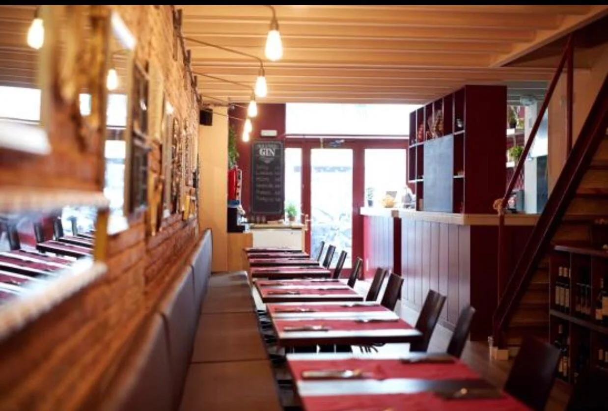 Interior de restaurante con mesas y sillas, decorado con madera, con iluminación cálida y una pizarra de bebidas al fondo.