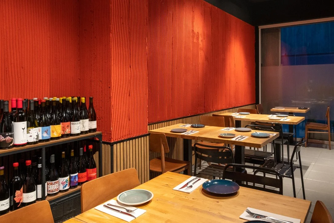 Interior de un restaurante con mesas preparadas con platos y cubiertos, y estantes con botellas de vino en una pared roja.