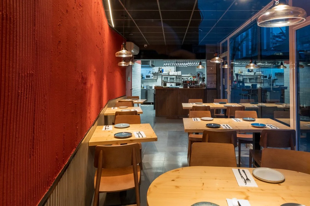 Interior de restaurante vacío con mesas de madera, platos y cubiertos, pared roja y vista a la cocina tras un mostrador.