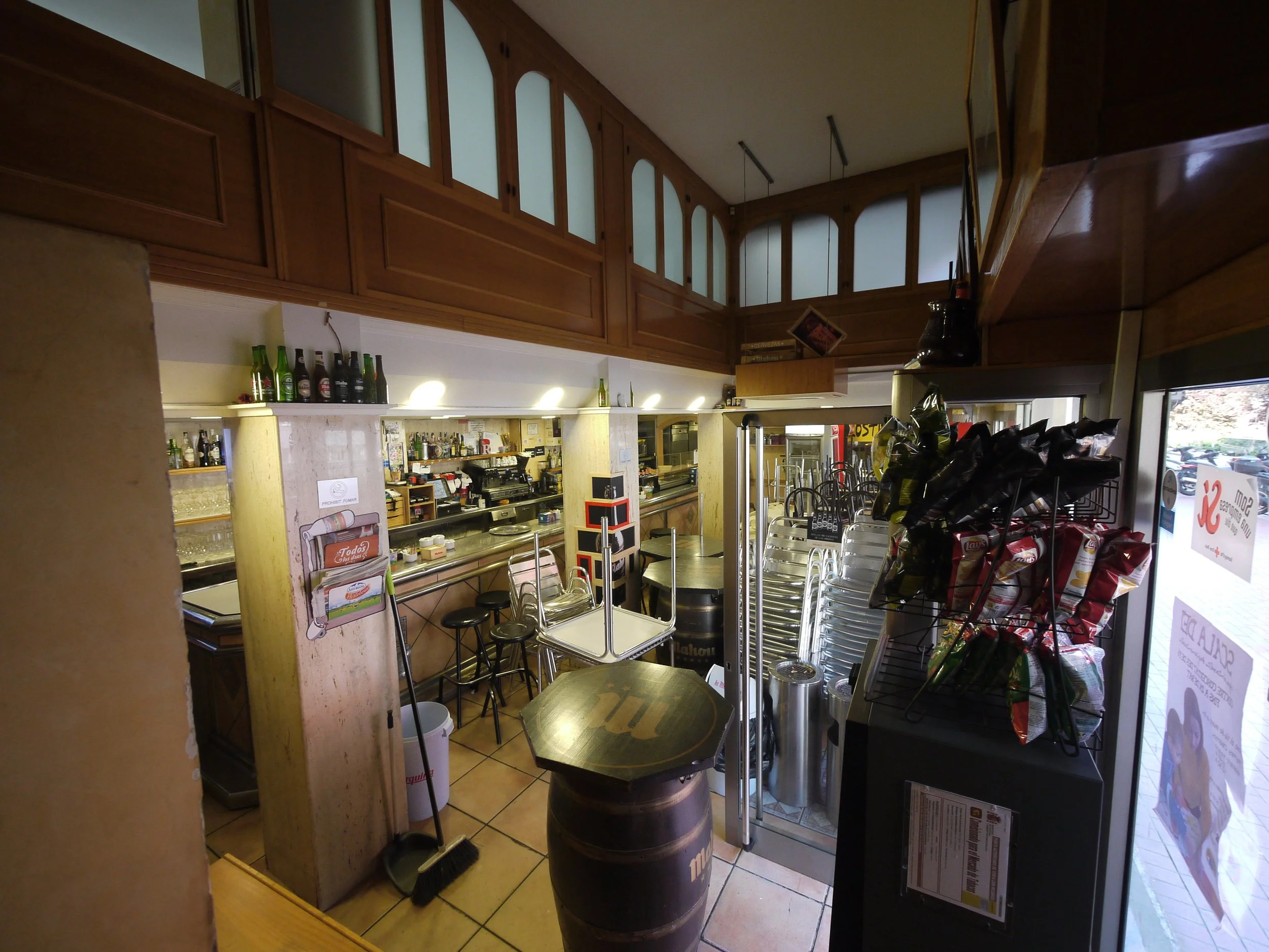 Interiores de un restaurante o bar con barra, mesas apiladas, mesas y sillas apiladas, estantes con botellas y snacks en exhibición.