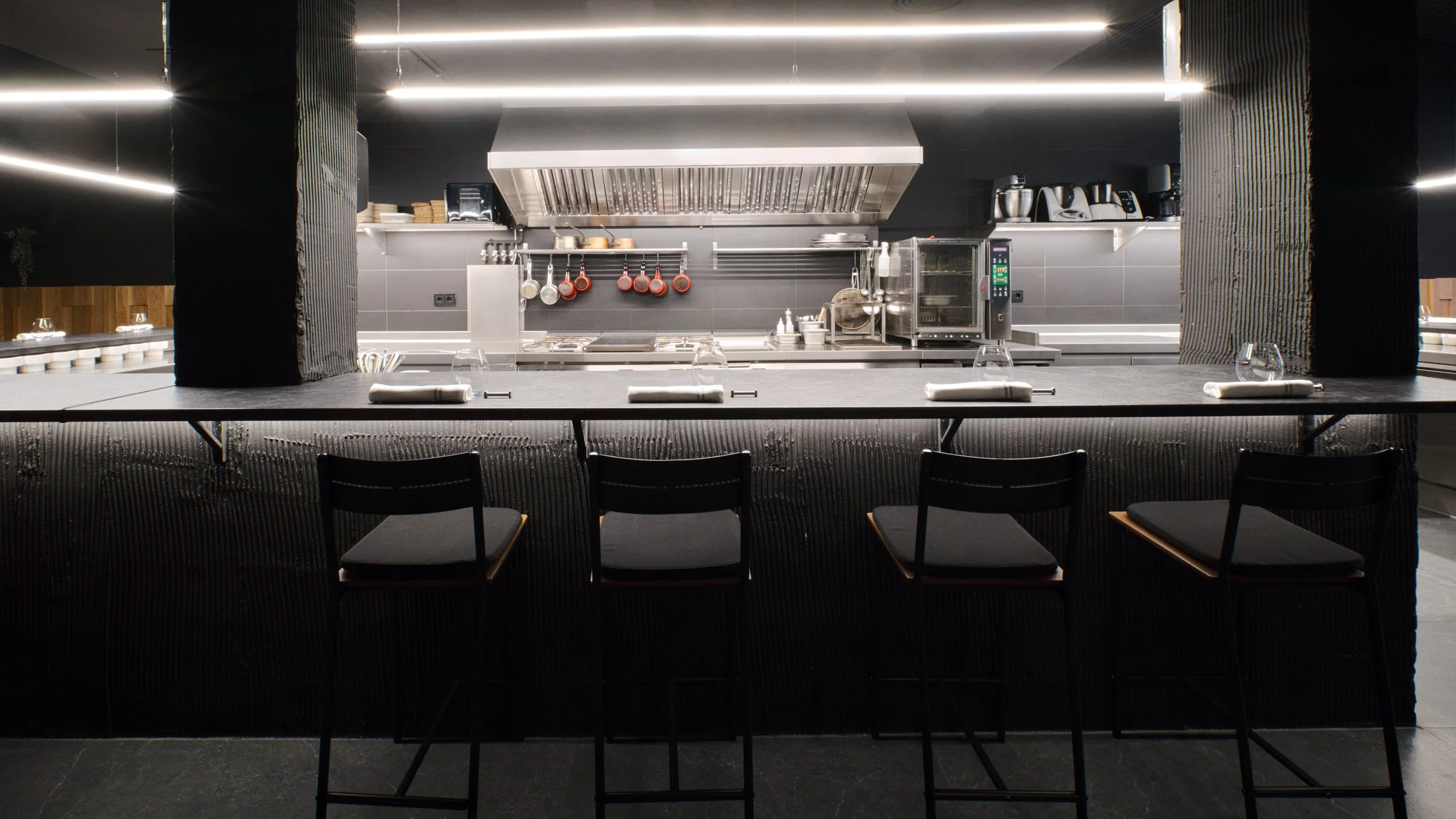 Mostrador de bar con sillas negras y un fondo de cocina moderna con estanterías, utensilios y electrodomésticos de acero inoxidable y negro.