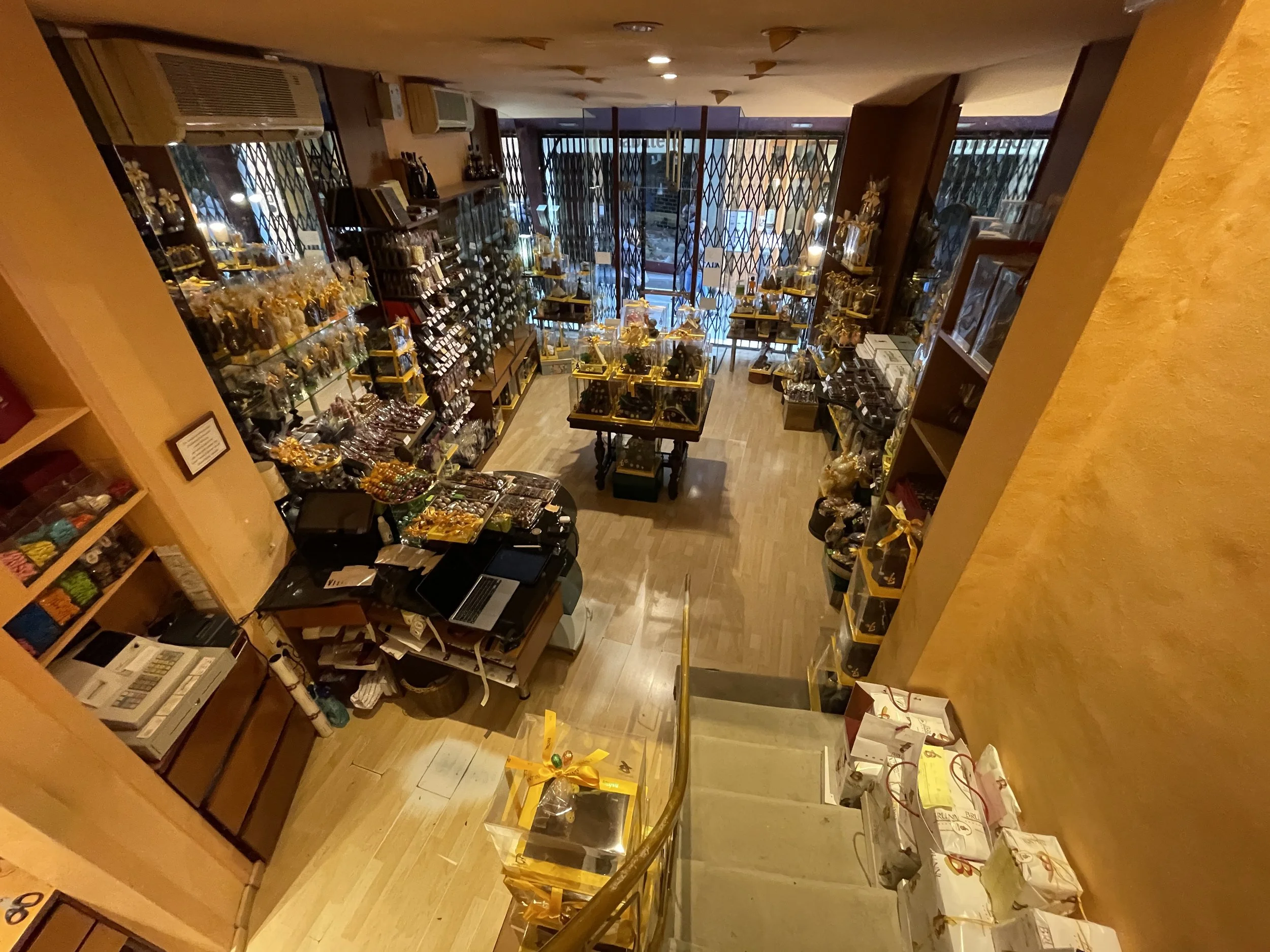 Vista desde arriba de una tienda con estantes llenos de productos, decoración y cajas, entrada con rejas de seguridad, y escalera con paquetes en la parte superior.