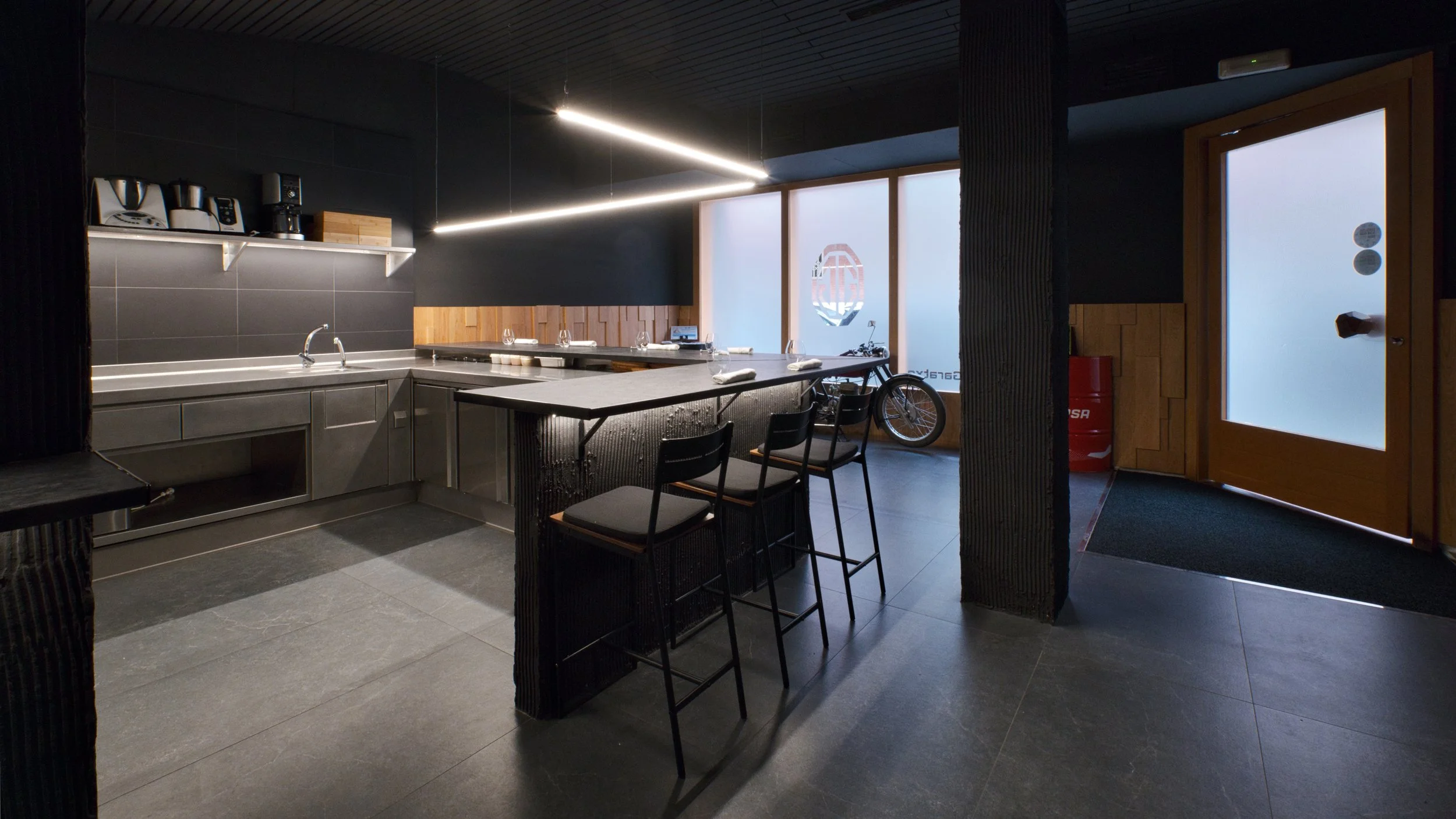 Cocina moderna con área de bar, encimeras de mármol, sillas altas, bicicleta junto a la ventana, puerta de madera con vidrio opaco.