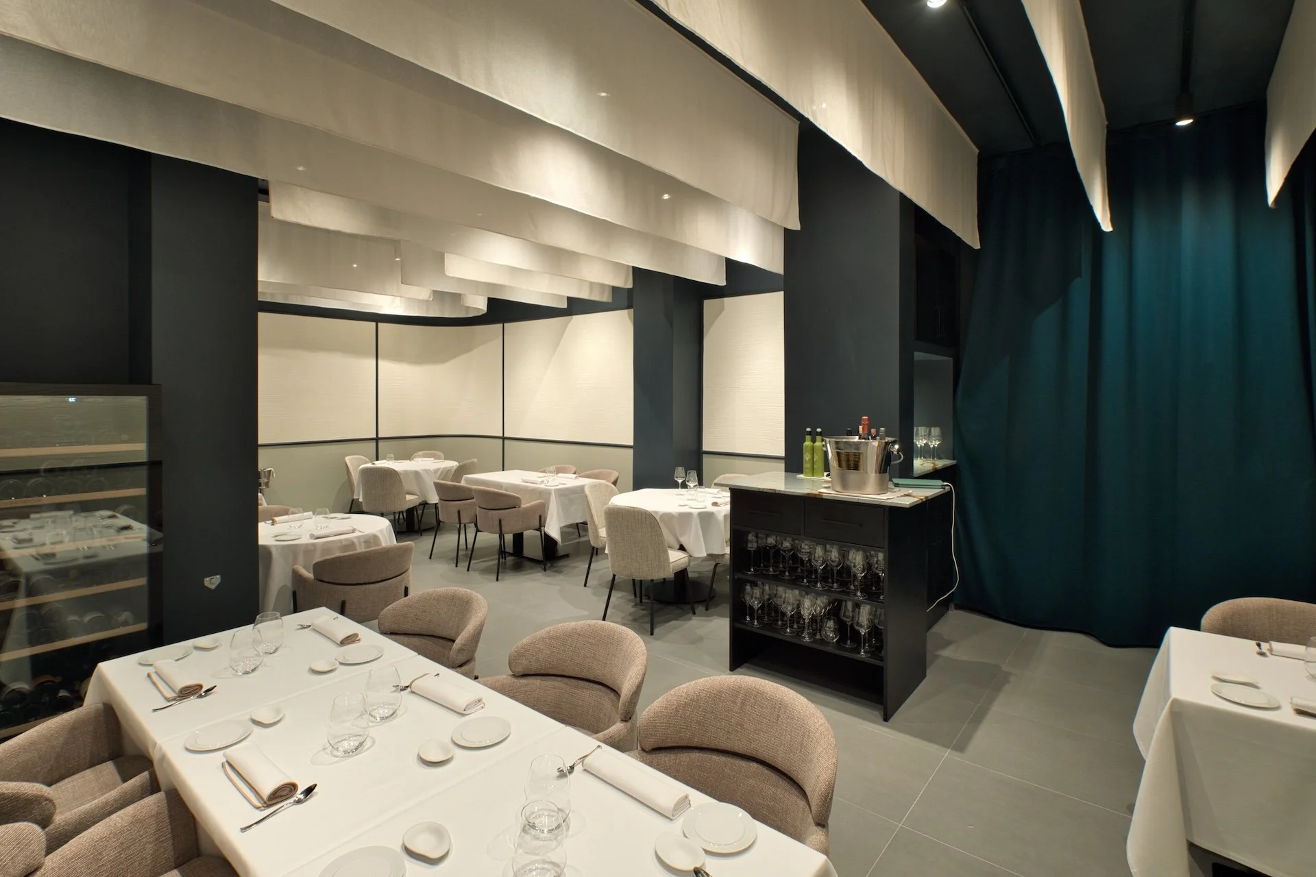 Interiores de un moderno restaurante con mesas cubiertas con manteles blancos, servilletas enrolladas, copas y platos, con sillas acolchadas en tonos beige y gris. Decoración minimalista con cortinas blancas colgantes en el techo y paredes de color oscuro, con un área de bar que tiene una nevera con botellas de vino y una barra con copas y una cubeta con botellas de vino y otras bebidas.