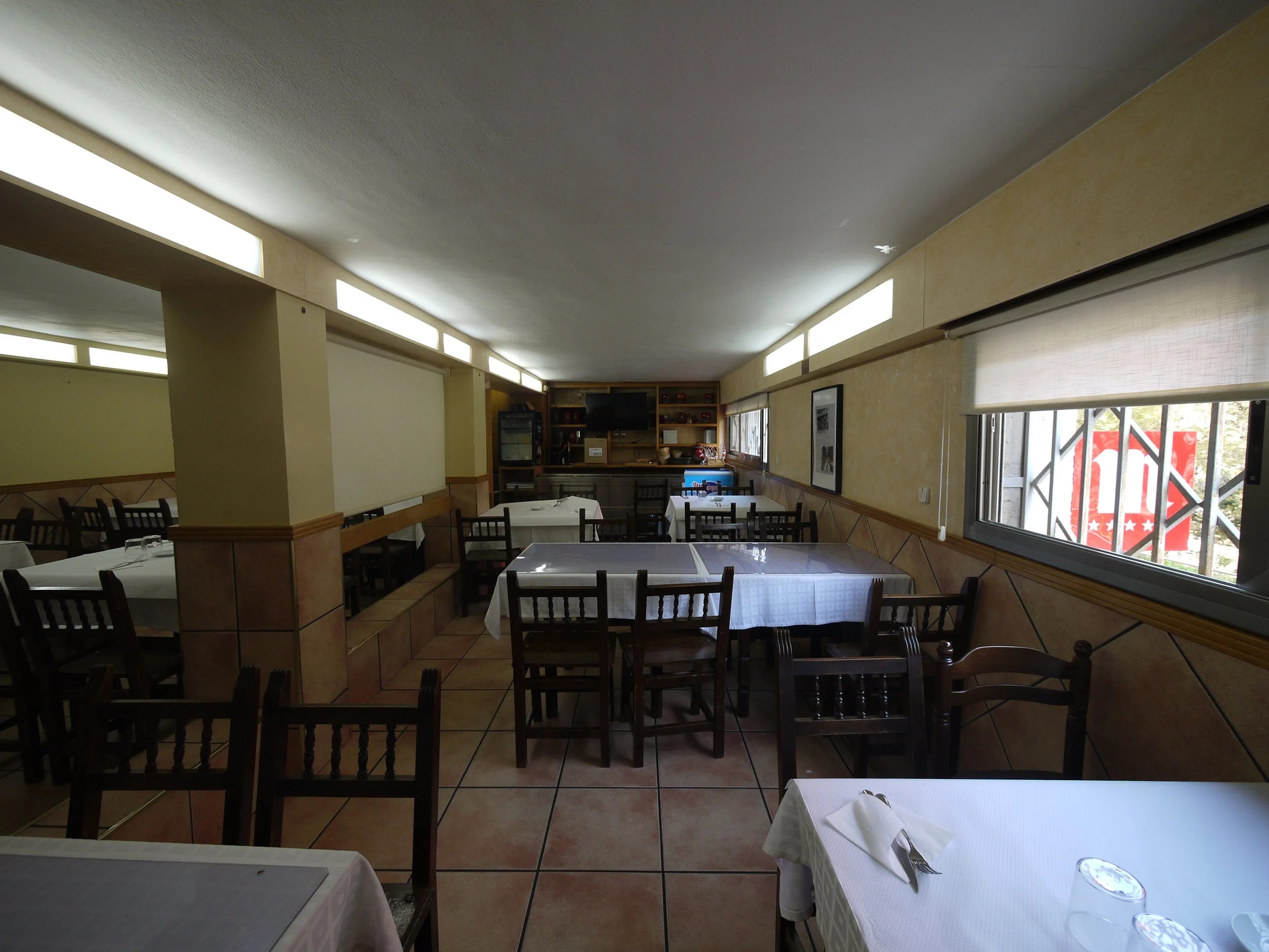 Interior de restaurante con mesas cubiertas con manteles blancos, sillas de madera, ventanas grandes y decoración sencilla.