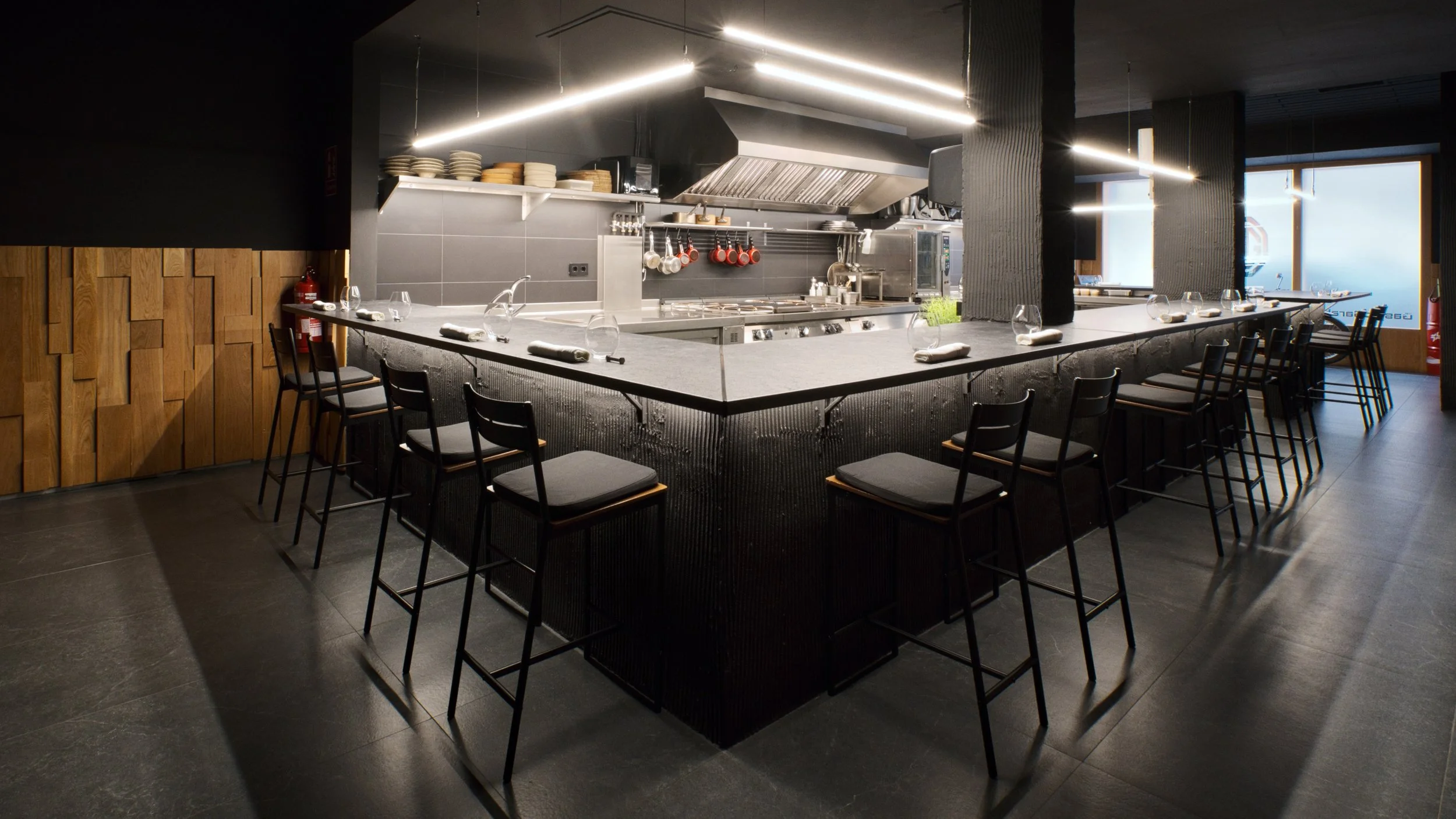 Mostrador de cocina en un restaurante elegante con sillas altas alrededor, utensilios y plantas decorativas, con estantes con platos y utensilios en la pared y ventanas al fondo.