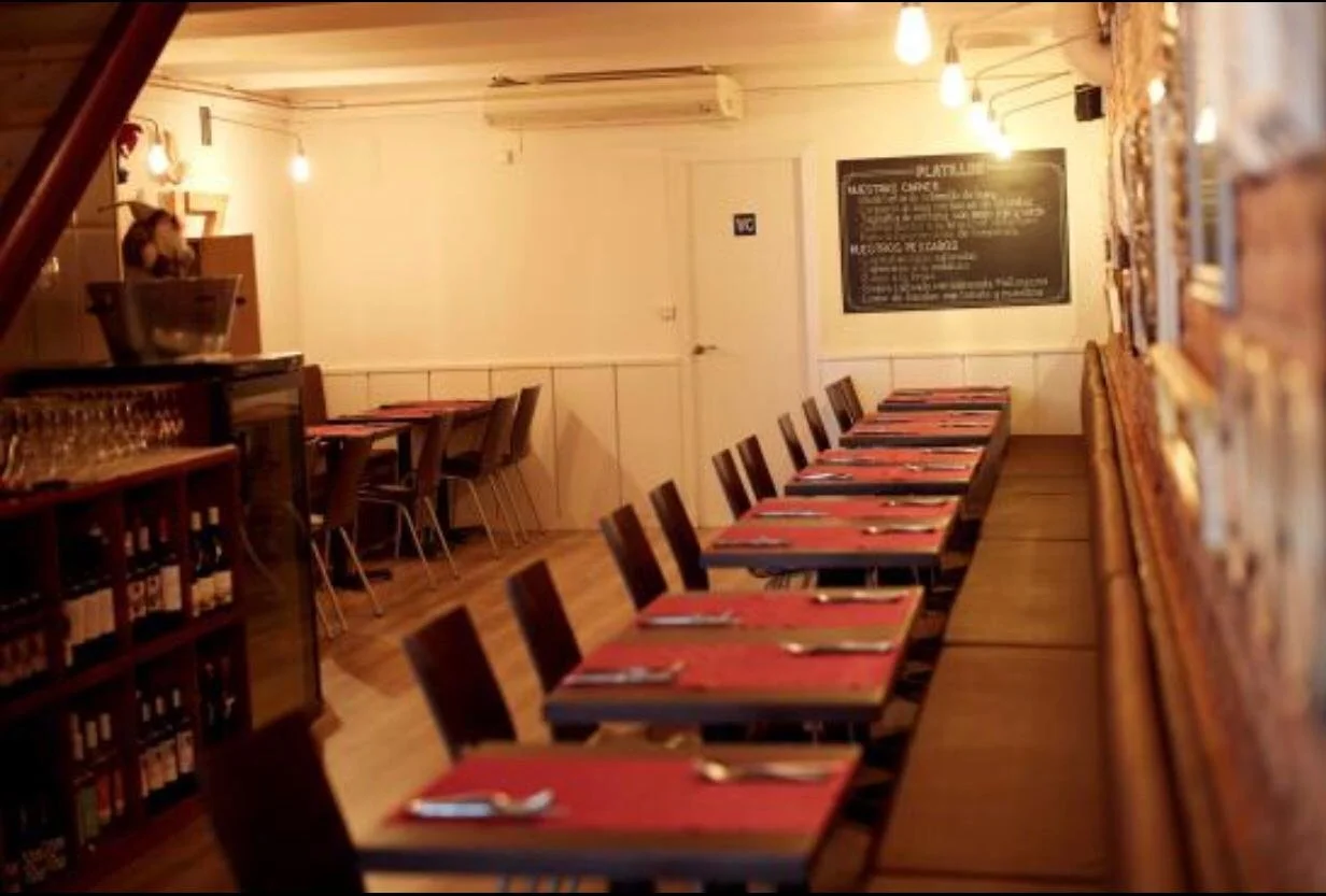 Interior de un restaurante con mesas arregladas y sillas, decoración cálida, pizarras con menús en la pared y iluminación tenue.