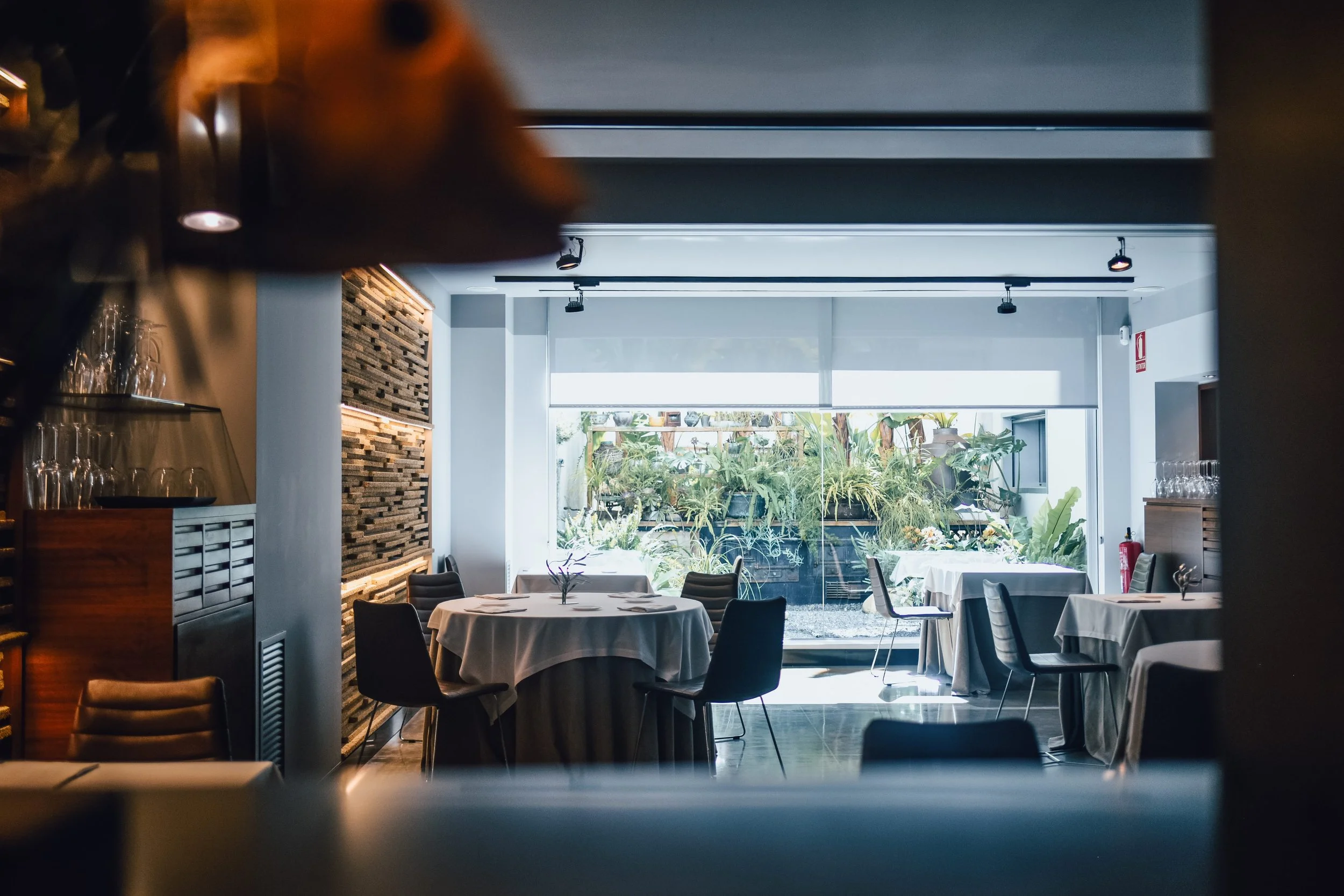 Interior de restaurante con mesas cubiertas con manteles blancos y sillas negras, vista a un jardín a través de un gran ventanal.