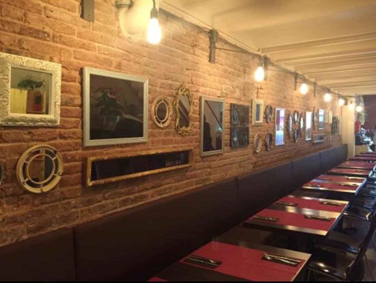 Interior de restaurante con pared de ladrillos vista, decorada con varios cuadros y espejos, mesas con manteles rojos y cubiertos, iluminación cálida con bombillas expuestas.