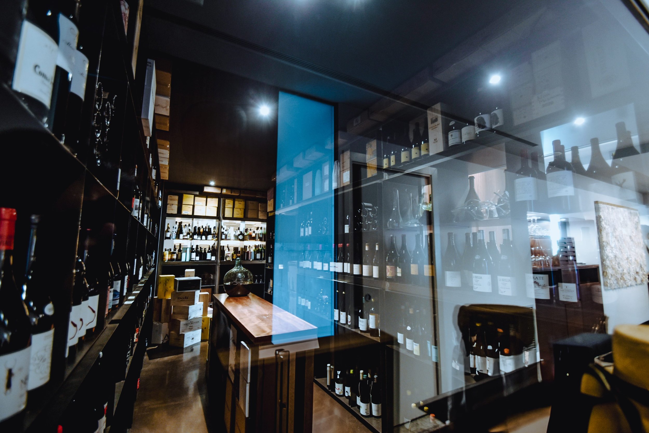 Interior de una tienda de vinos con estanterías llenas de botellas de vino, vista reflejada en un vidrio.