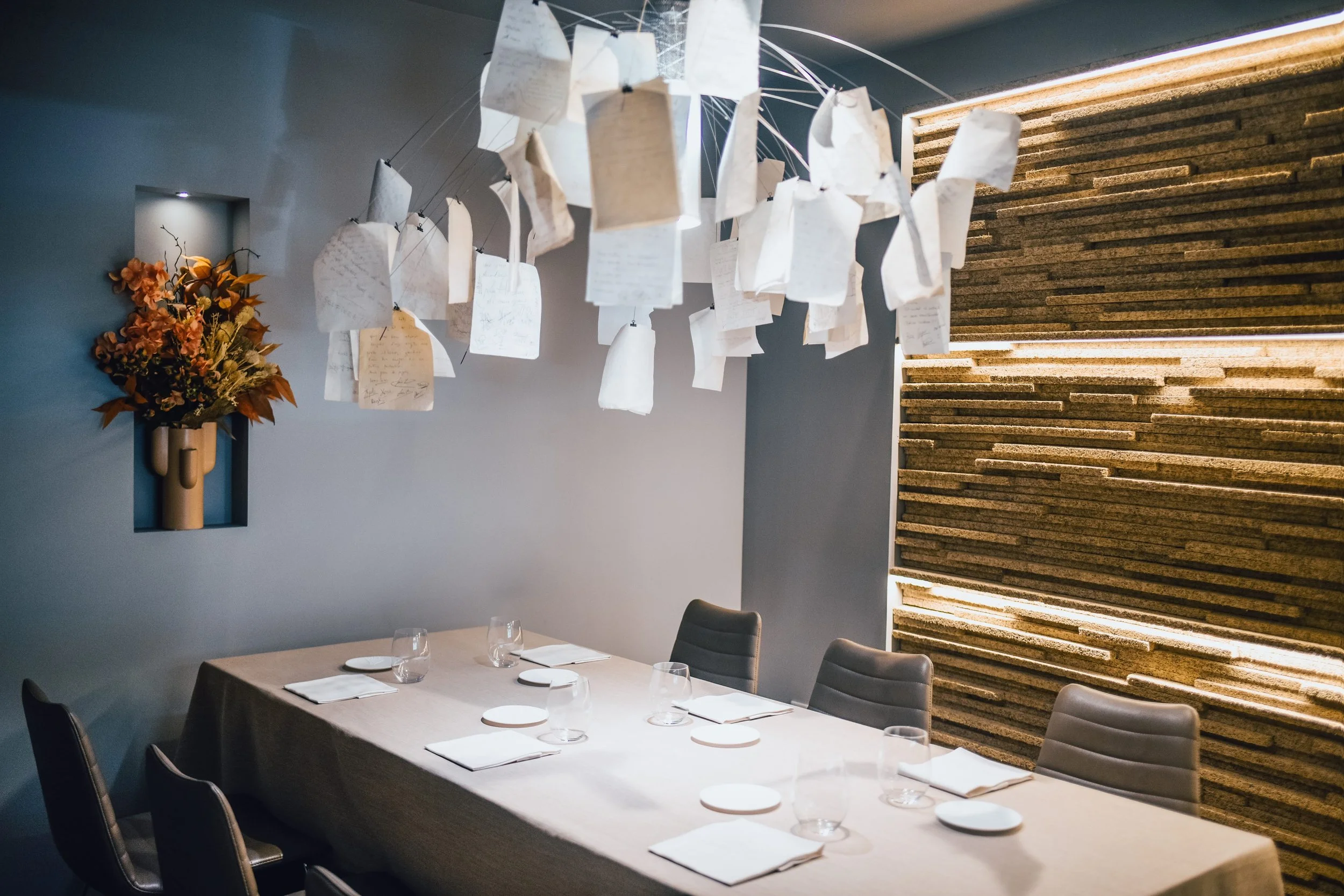 Sala de comedor moderna con mesa rectangular, sillas negras y lugar preparado con copas, platos y servilletas. Decoración en la pared con una repisa de flores y una instalación artística con papeles colgantes encima de la mesa.