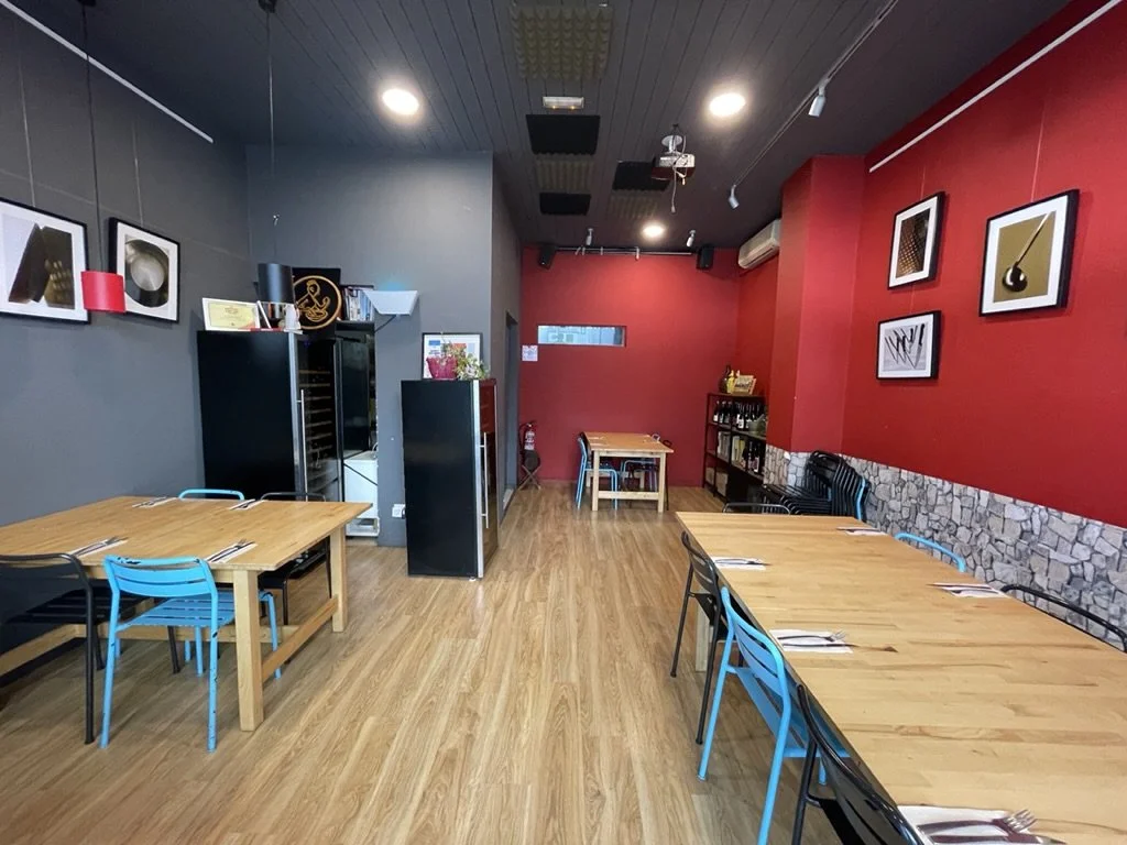 Interior de un restaurante con paredes grises y rojas, mesas de madera, sillas negras y azules, decoraciones modernas en las paredes y una estantería con botellas y objetos decorativos.