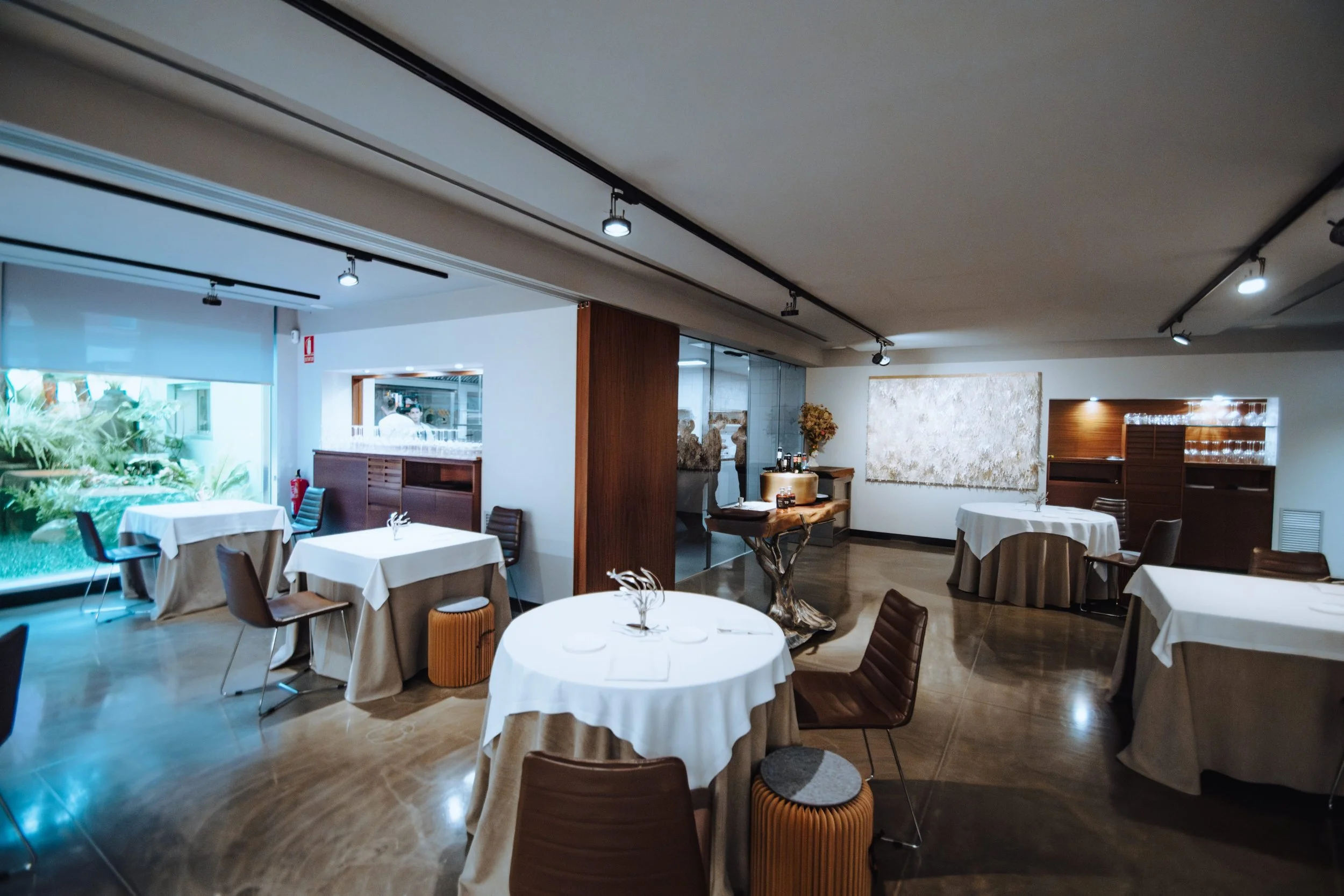 Restaurante elegante con mesas cubiertas con manteles blancos y sillas de madera, decoraciones con plantas y obras de arte en las paredes, y una vista a un área con vegetación exterior