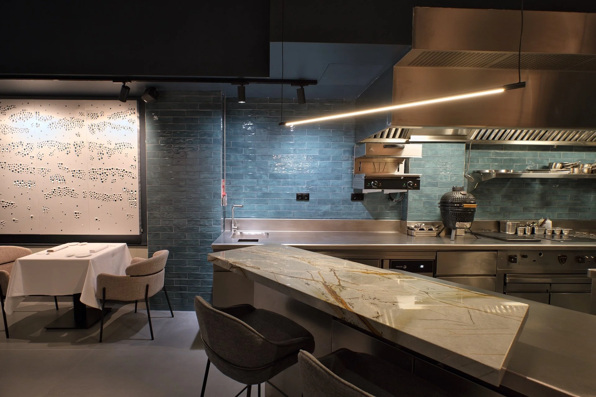 Cocina moderna con paredes de azulejos azules, barra de mármol y área de comedor con sillas y mesa con mantel blanco.