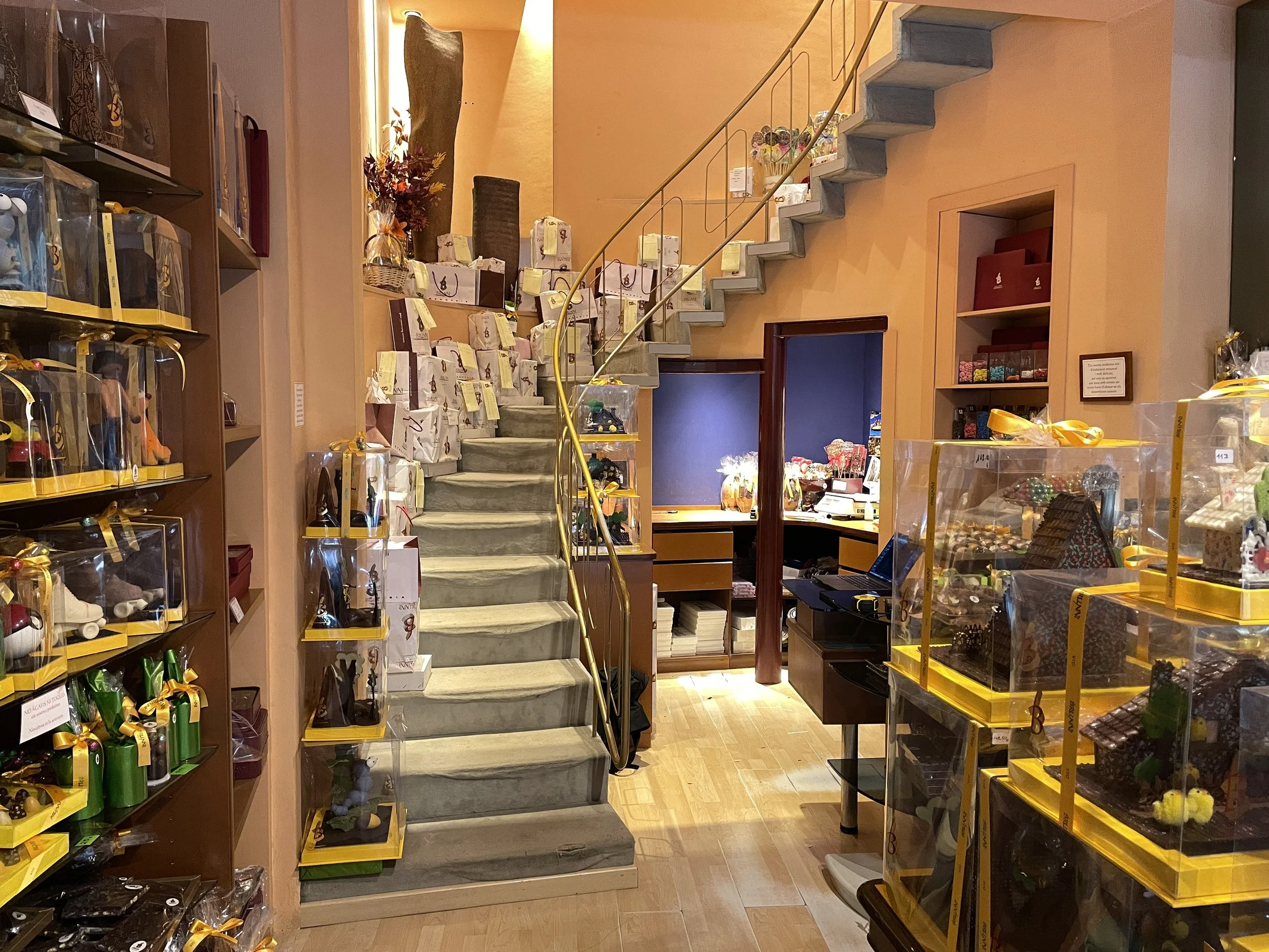 Interior de una tienda decorada con figuras y regalos en exhibición, incluyendo muchas cajas con artículos envueltos y decorativos, y una escalera que lleva a un segundo nivel.