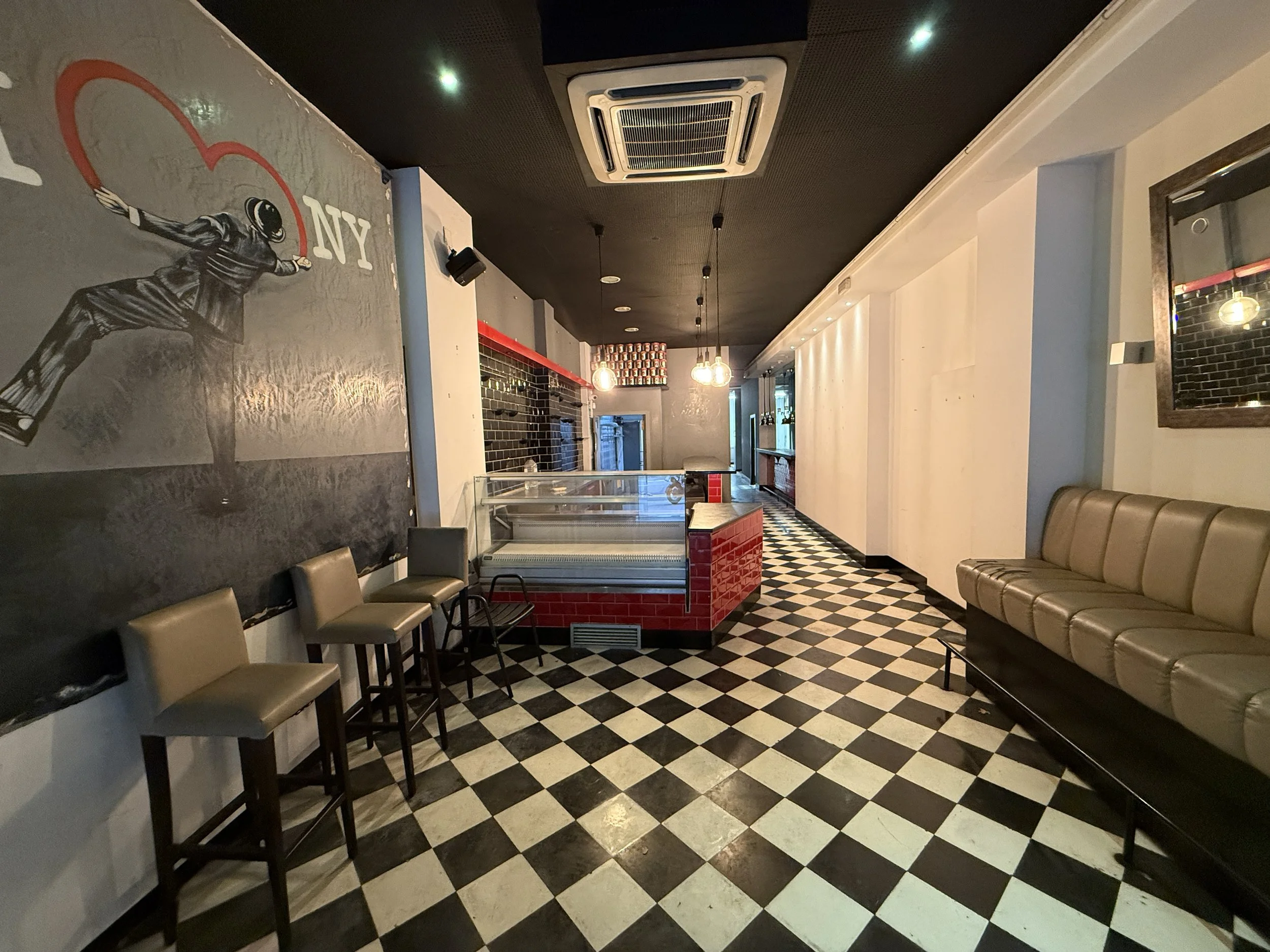 interior de un restaurante con paredes blancas, una banqueta beige en el lado derecho, cuatro sillas grises en el lado izquierdo y un mostrador con rejas negras y un frente de azulejos rojos, suelo de baldosas negras y blancas en patrón de checkerboard, un mural en la pared izquierda y un espejo en la pared derecha