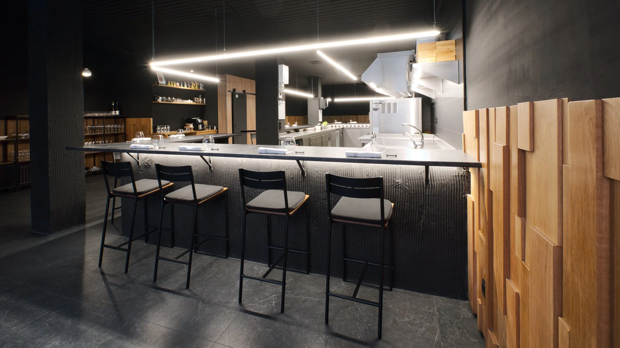 Barra de restaurante moderna con cinco sillas lista para comensales, cocina al fondo con electrodomésticos de acero inoxidable y estantes con utensilios.