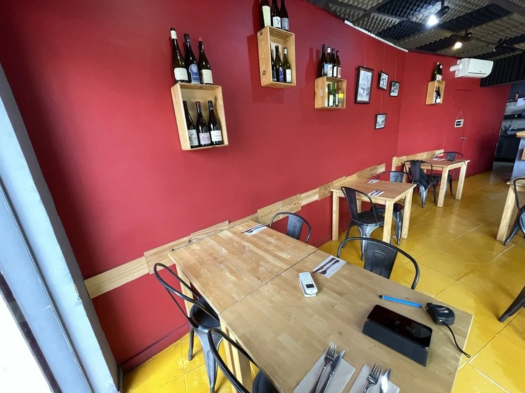 Mesa de madera con cubiertos y electrodomésticos en un restaurante con paredes rojas y decoración de botellas de vino en estantes de madera.