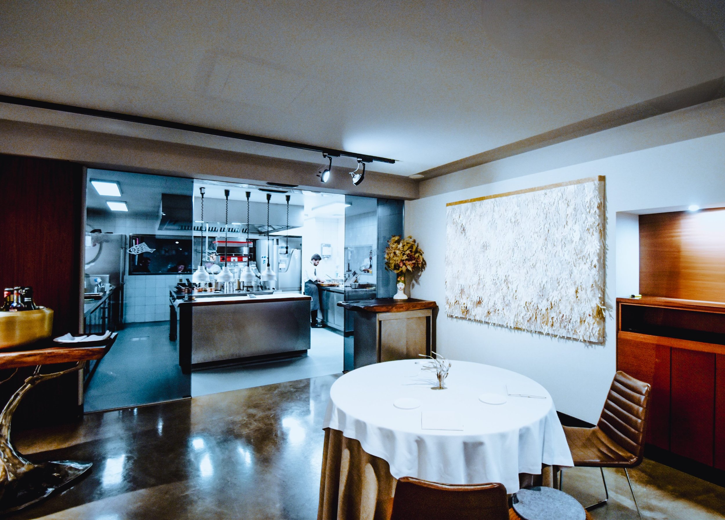 Vista de un comedor en un restaurante elegante con mesa redonda, sillas de madera y mantel blanco, decoraciones florales y una pared con arte, con una cocina abierta en el fondo y cocina en funcionamiento.