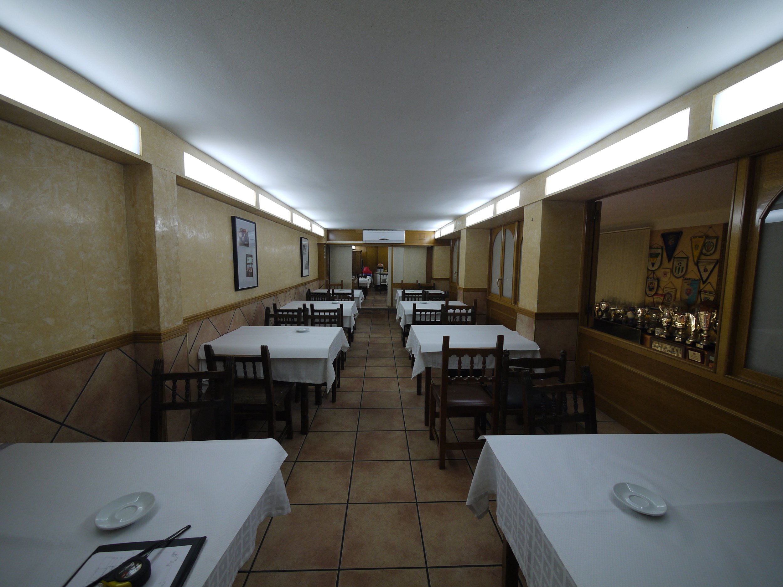 Salón de restaurante vacío con mesas cubiertas con manteles blancos y sillas de madera. La pared de fondo tiene decoraciones y una vitrina con trofeos.