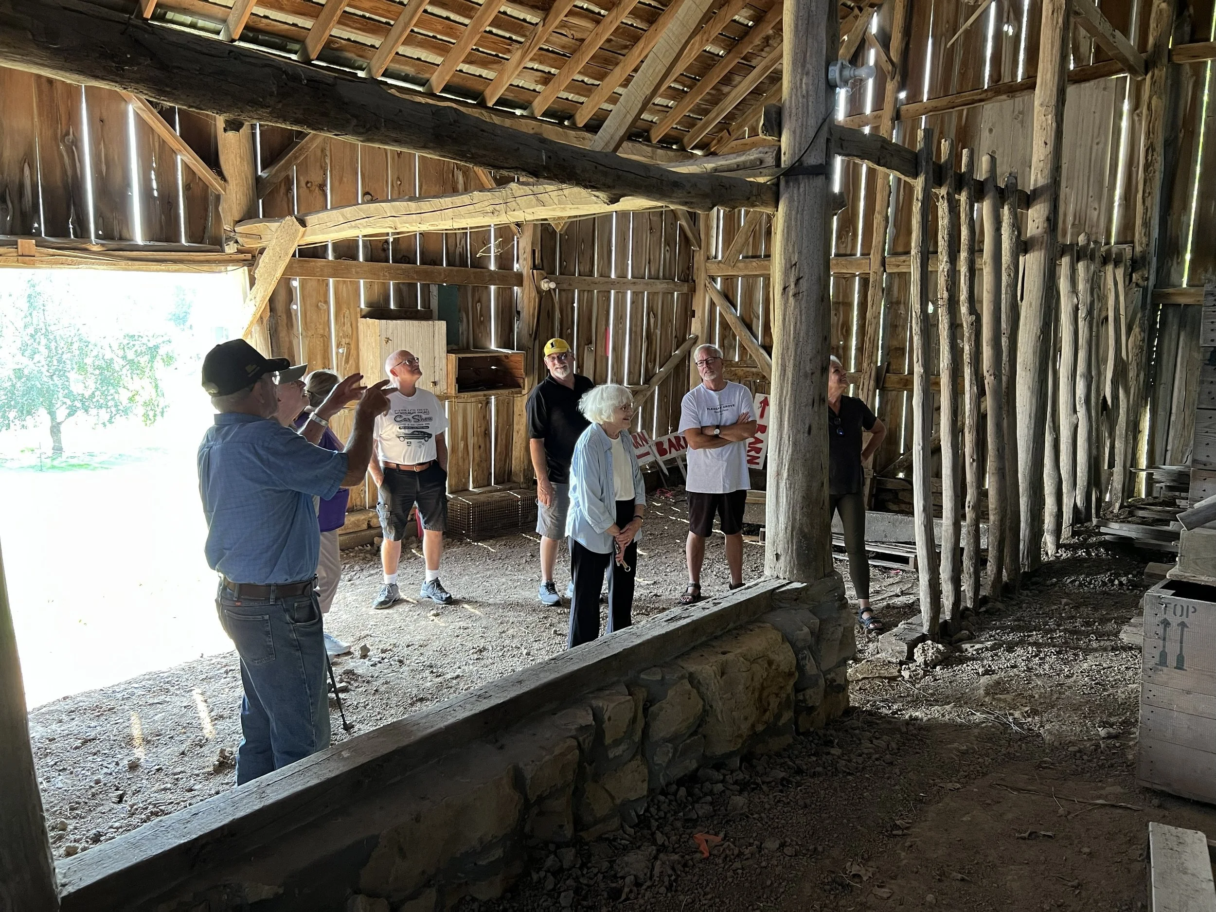 A group of people is inside a rustic wooden barn, listening to a person in a light blue shirt and black cap give a presentation or speech. The barn has exposed wooden beams and walls, with sunlight streaming in from the left side.