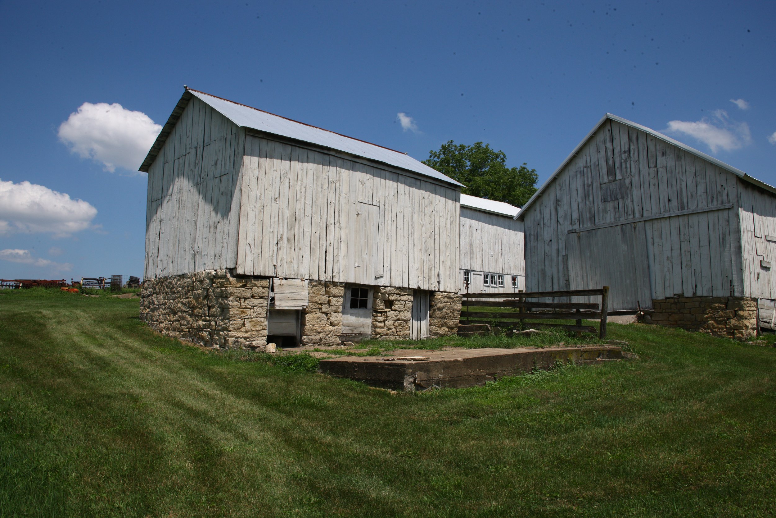 Two old white wooden barns with stone foundations on a grassy field under a blue sky with scattered clouds.