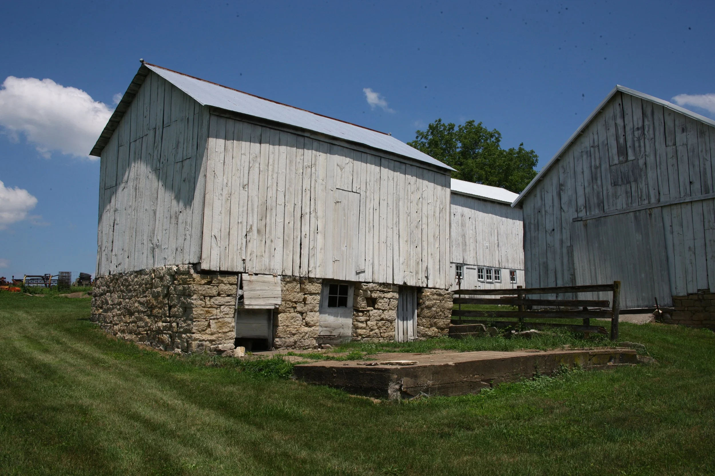 Two white wooden barns on a grassy field under a blue sky with a few clouds, with one barn on stone foundation and small windows.