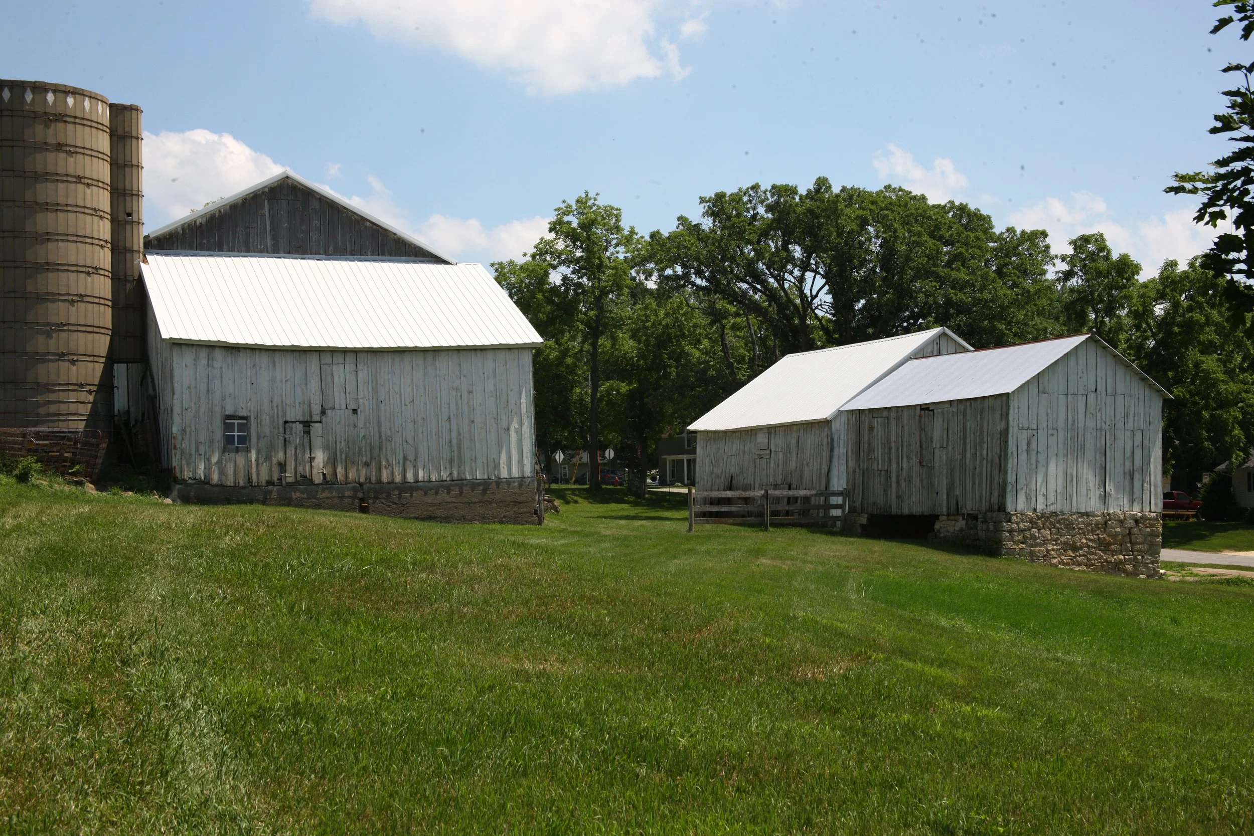 Two old wooden barns with white metal roofs in a grassy field, trees in the background, and a partly cloudy sky above.