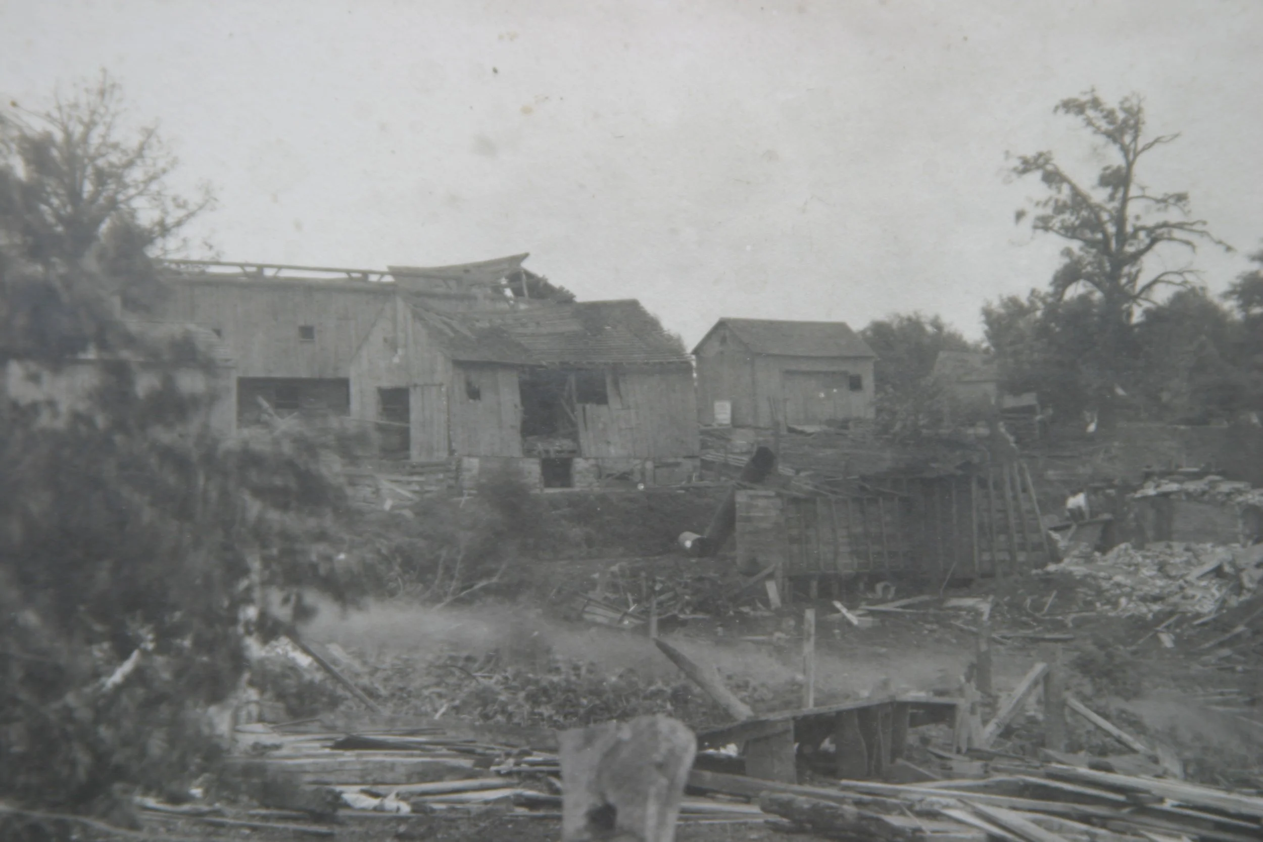 Black and white photo of damaged wooden houses, with debris and fallen structures in the foreground and trees in the background.