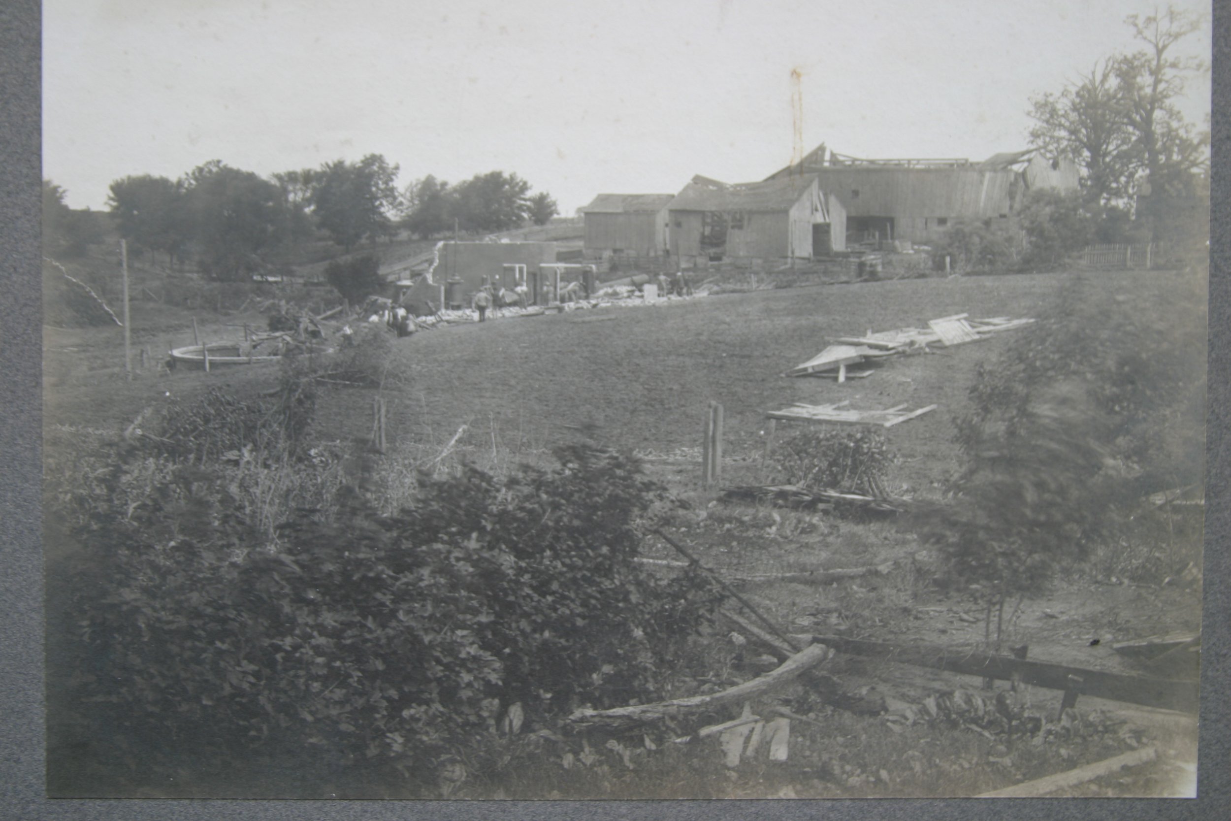 Black and white photo of a damaged farm or rural scene with debris, fallen structures, and a partially collapsed building in the background.