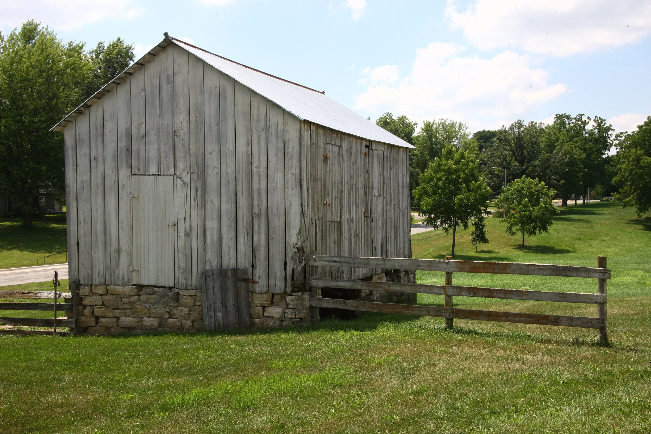 An old weathered wooden barn with a stone foundation, surrounded by green grass and trees on a sunny day.