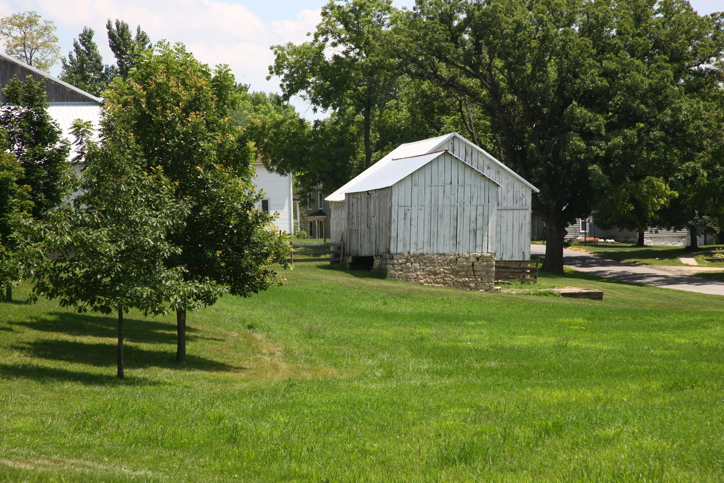 A small weathered white barn with a metal roof, elevated on stone foundation, surrounded by green grass and trees, in a quiet residential area.