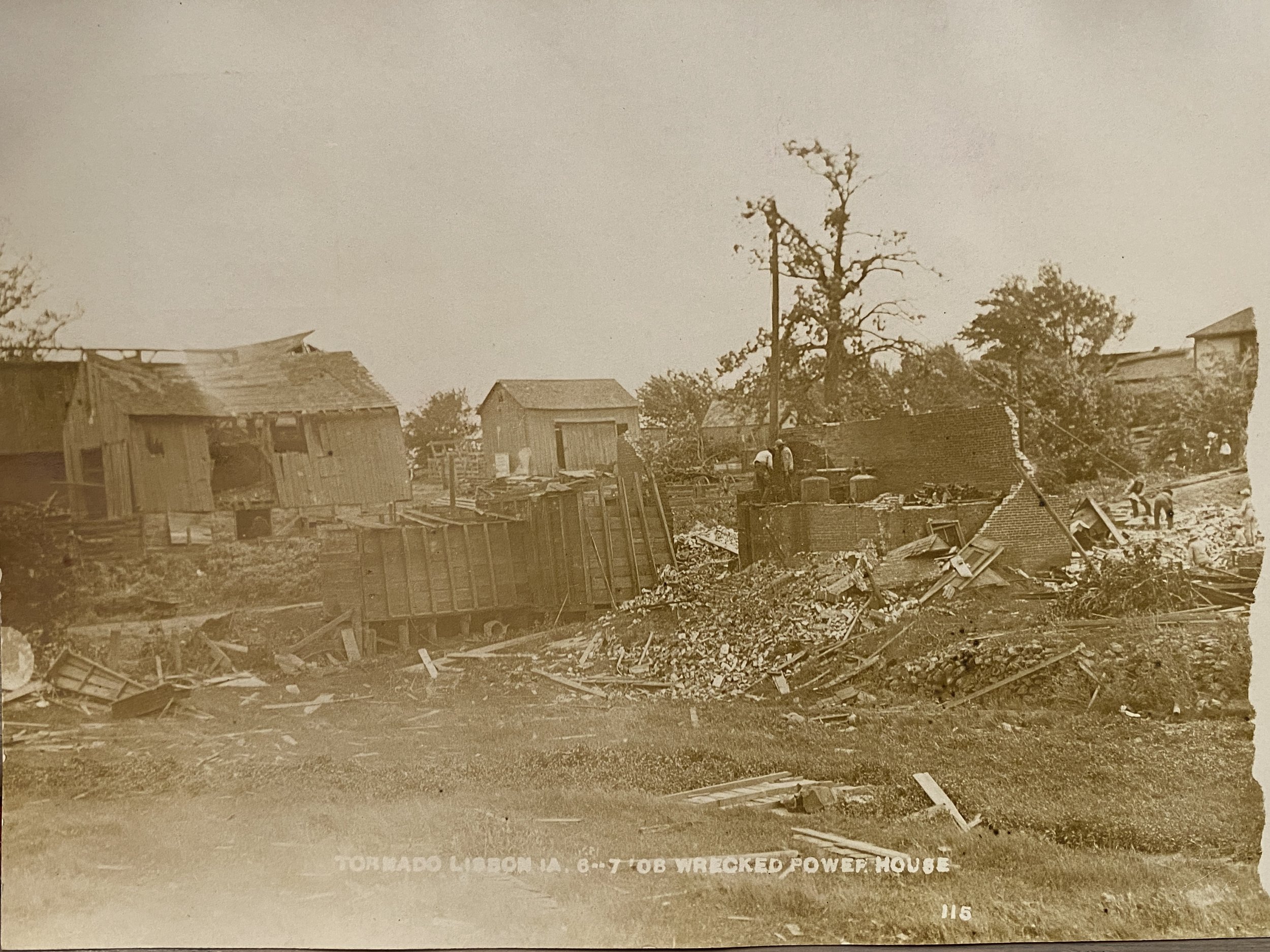 Black and white photo of a damaged building with debris scattered on the ground, a fallen utility pole, and workers in the background, likely after a tornado or storm.
