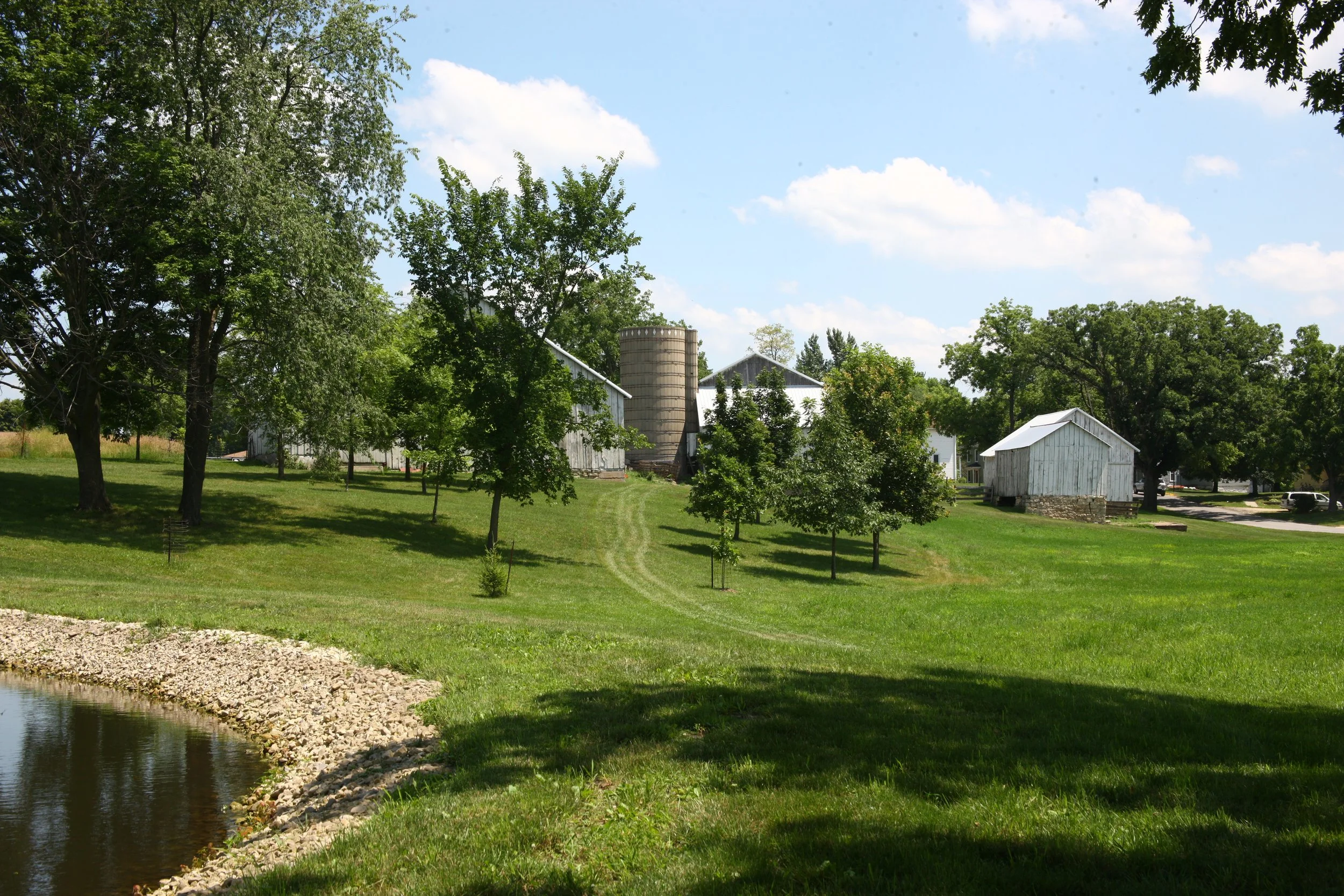 A peaceful rural scene featuring a pond with a rocky shoreline on the left, lush green grass, several trees, and white farm buildings with silos in the background under a partly cloudy blue sky.