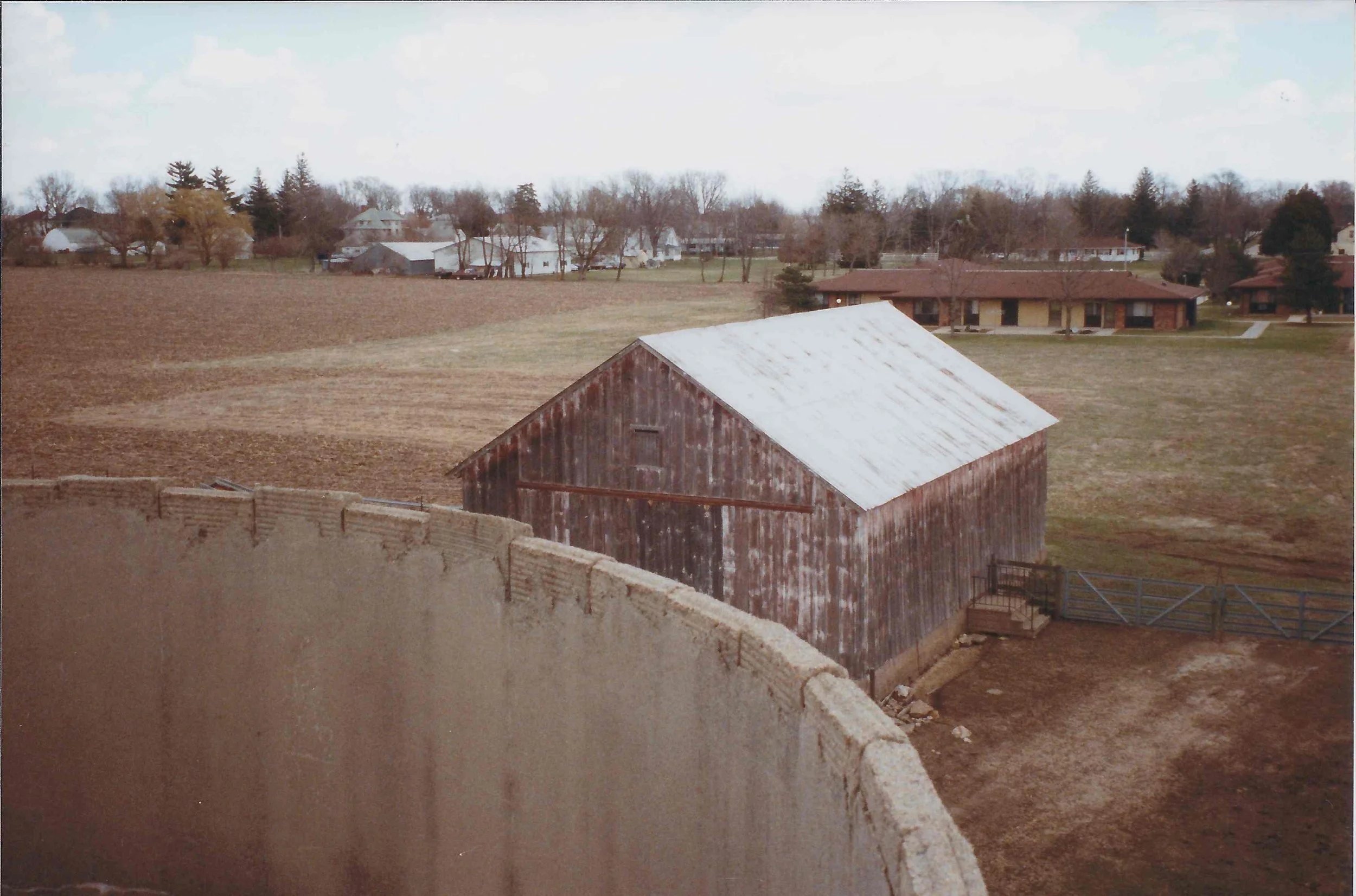 A rustic barn with weathered red wood siding and a white metal roof, situated in an open field with a concrete wall in the foreground.