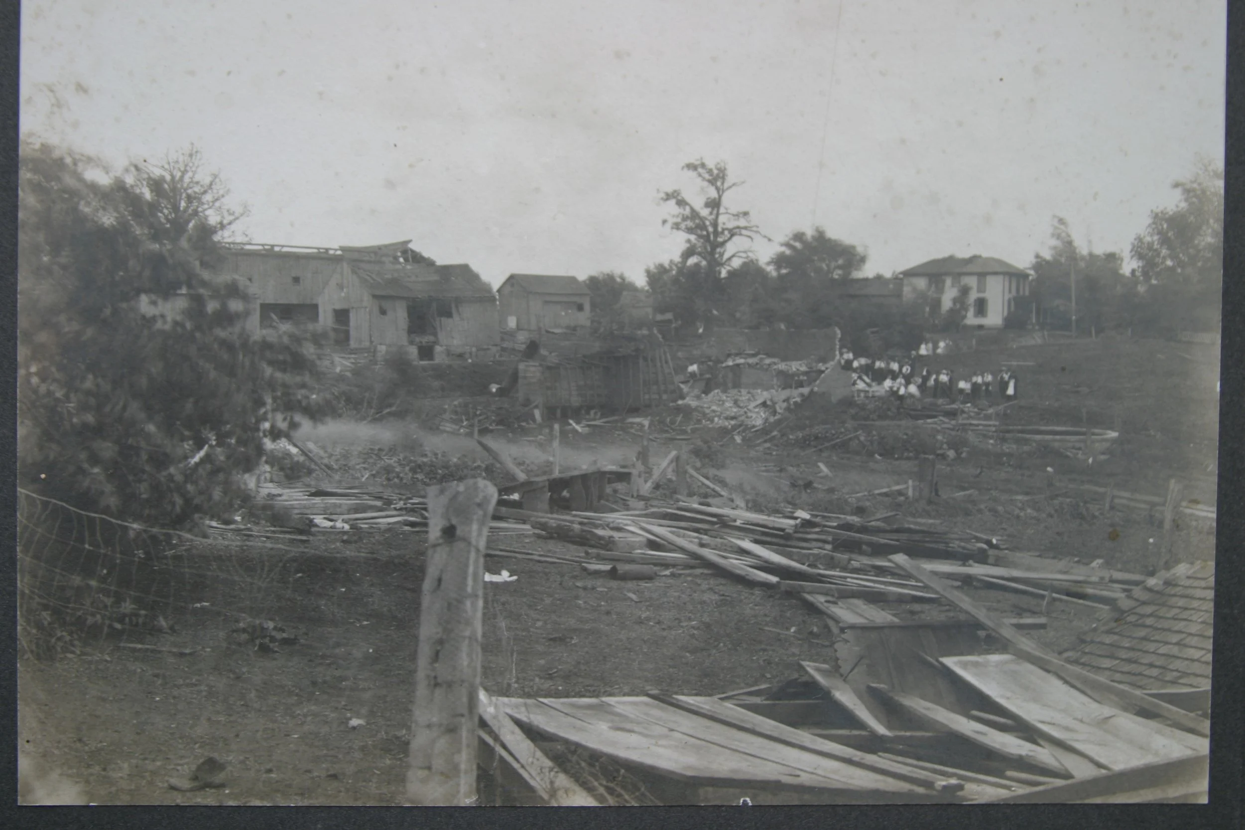 Black and white photograph of a landscape with damaged houses, debris, and a group of people in the distance, possibly children.