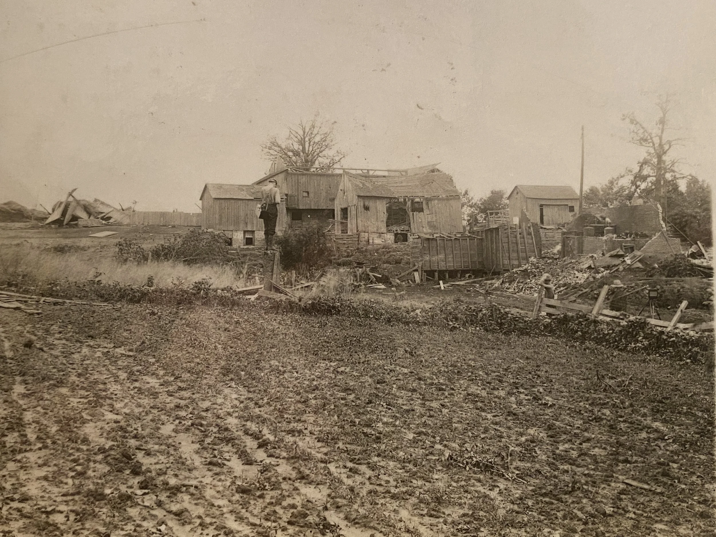 A vintage black and white photo of a rural area with dilapidated wooden houses, debris, and a man standing on a small bridge or platform near the houses. The ground in the foreground appears to be plowed or muddy.