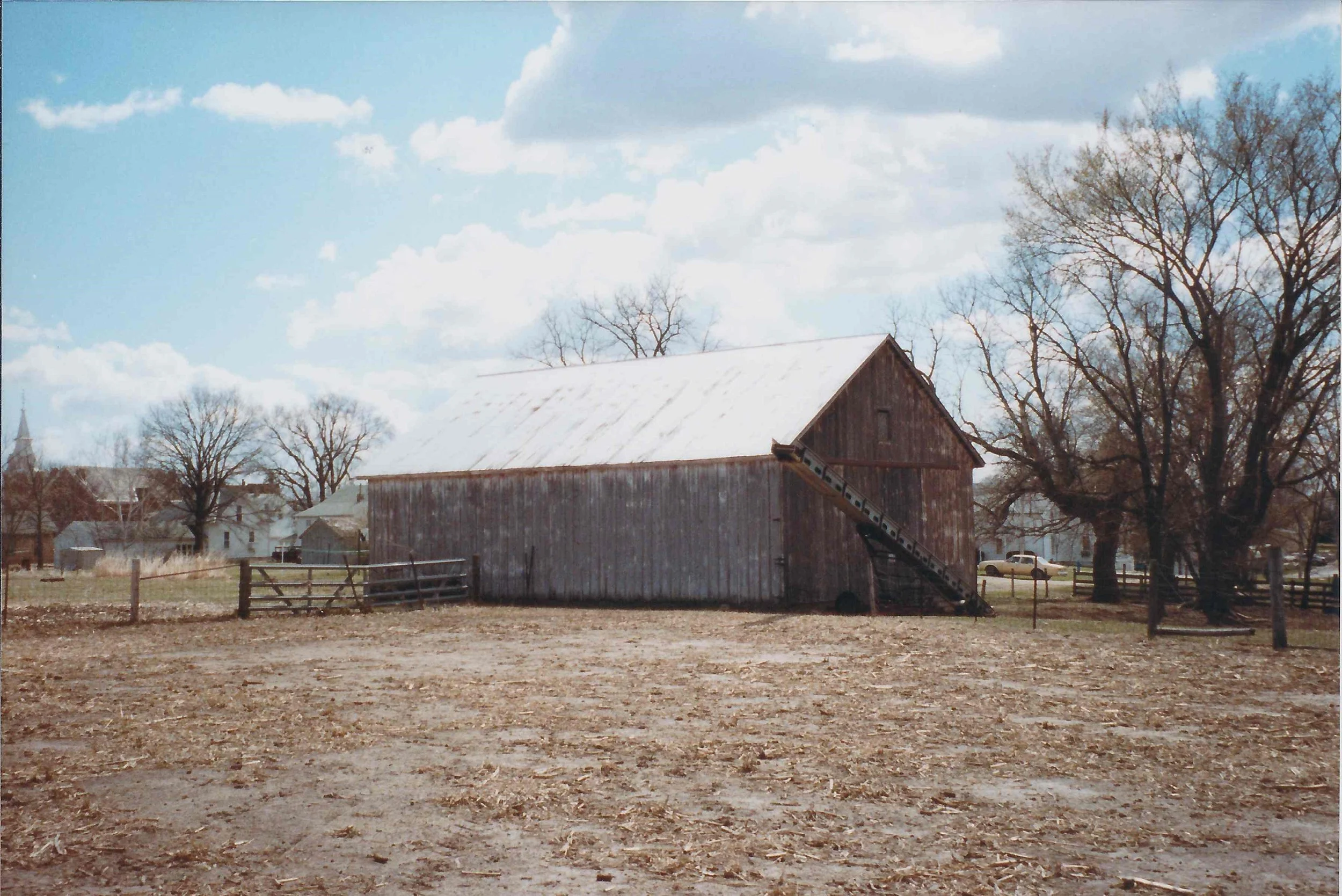 An old wooden barn with a snow-covered roof and an outdoor staircase on the side, standing in a rural setting with bare trees and a partly cloudy sky in the background.