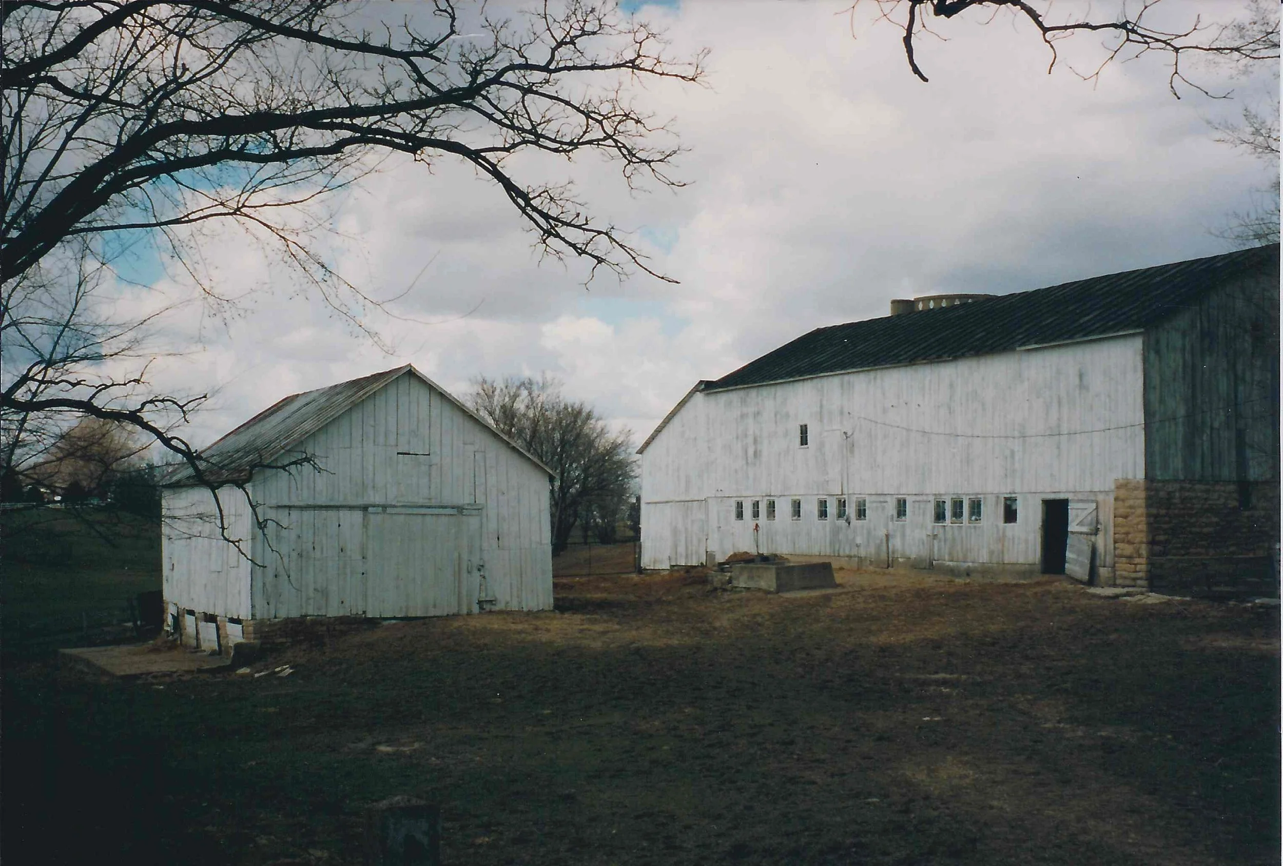 Two white wooden barns on a farm with cloudy sky and leafless trees.