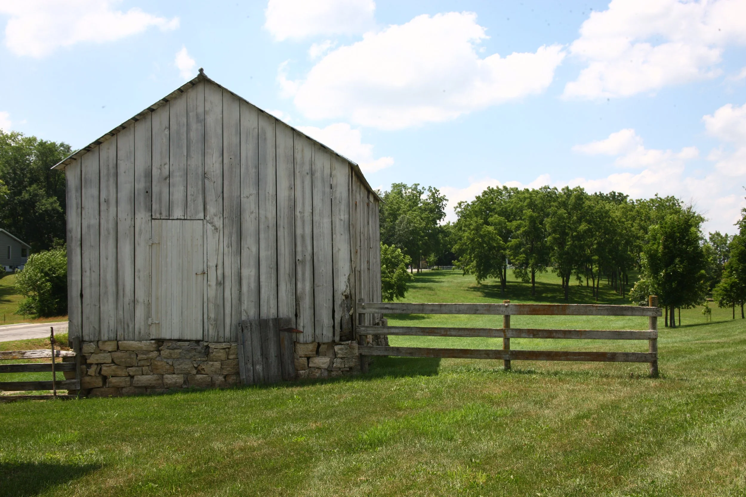 Old wooden barn with a stone foundation in a rural setting, with green grass, trees, and a partly cloudy sky