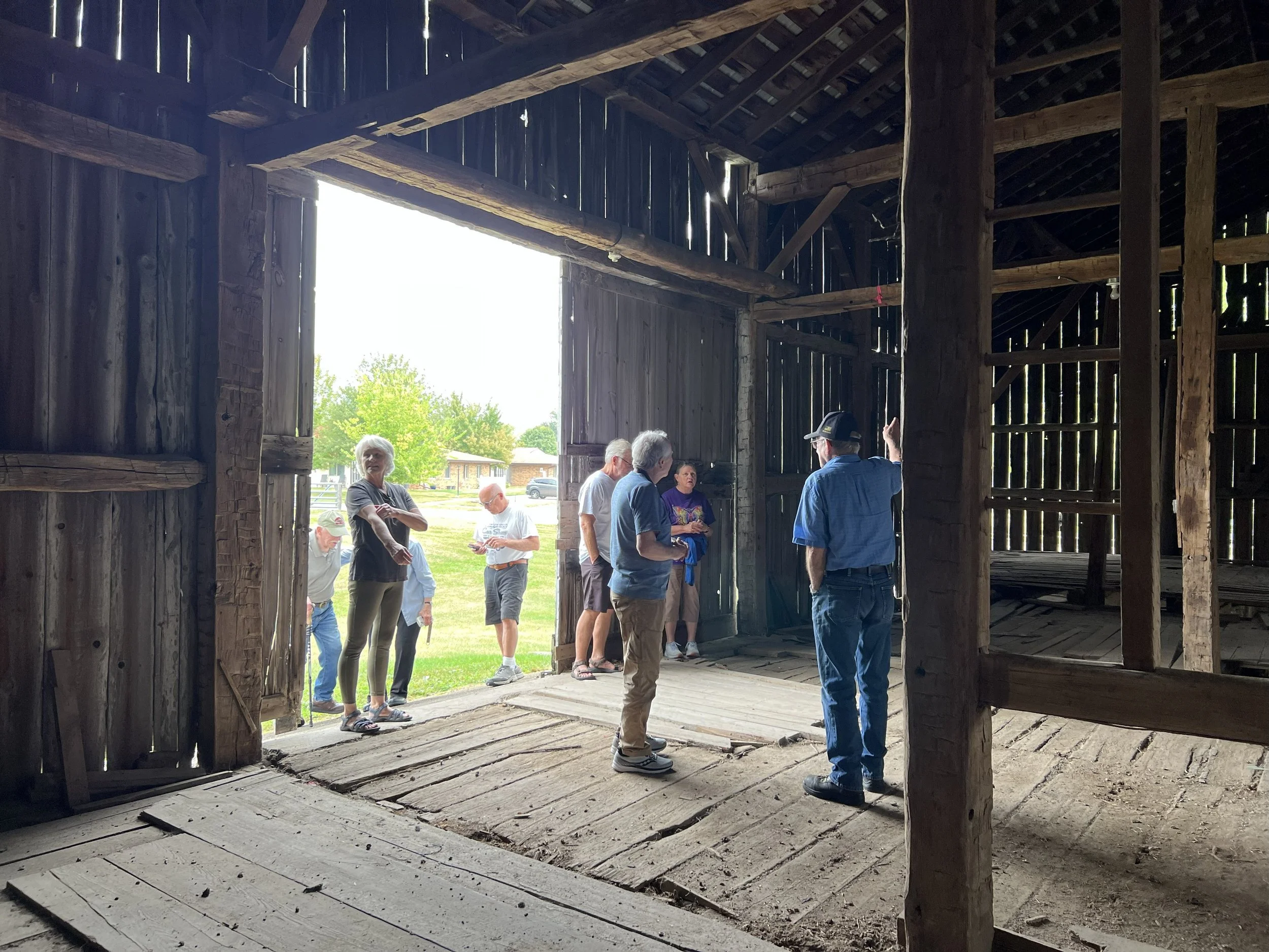 A group of people gathered inside an old wooden barn with weathered walls and exposed beams, some standing near the open doorway looking outside, where green trees and a building are visible.