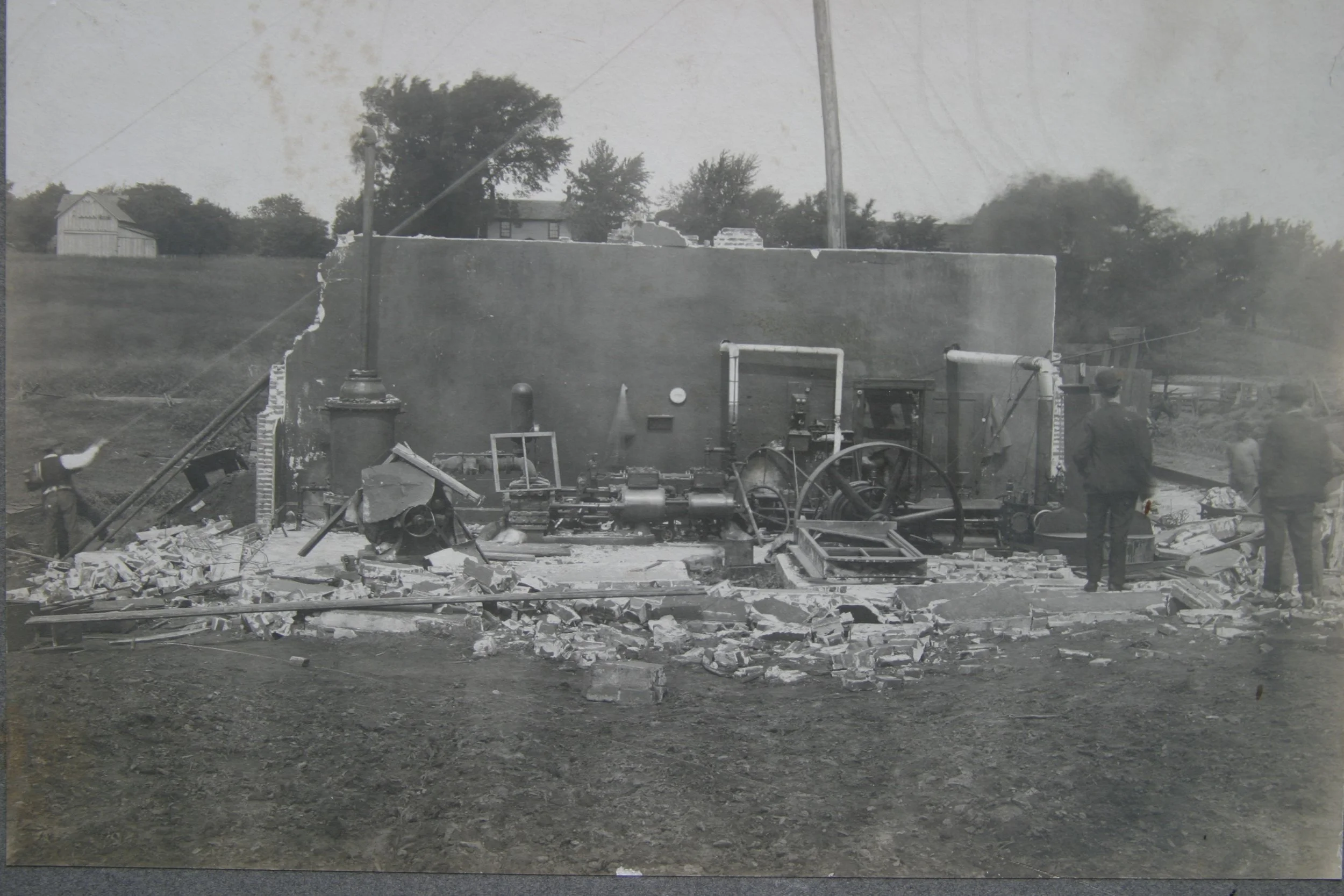 Black and white historic photo of a demolished building with debris, debris, bricks, and remnants of machinery scattered on the ground, and several men inspecting the site.