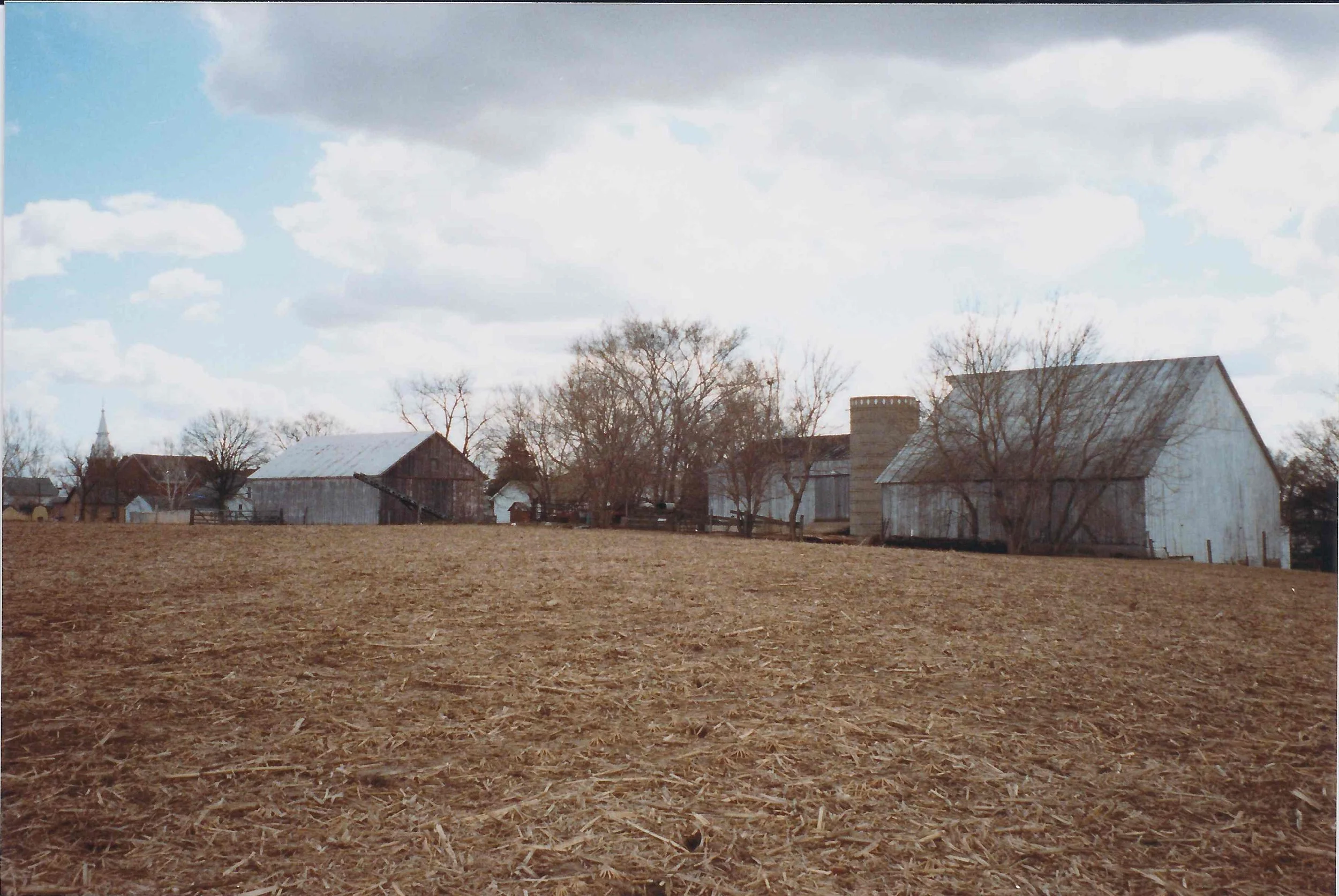 A rural scene with a plowed field in the foreground and several old wooden barns and houses in the background under a partly cloudy sky.