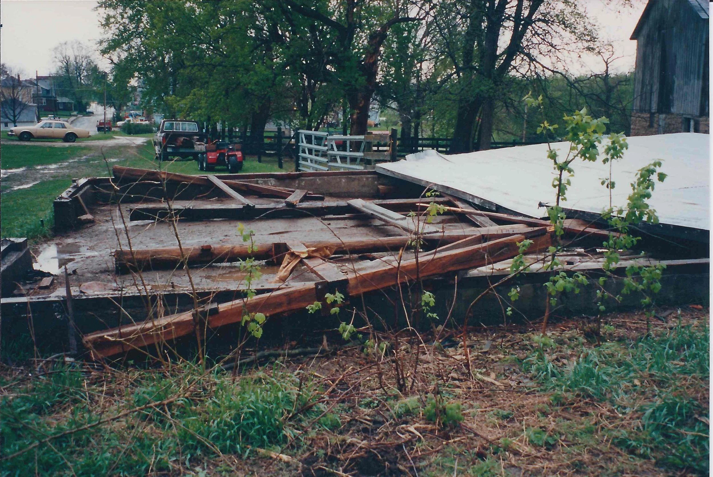 Remnants of a collapsed structure, possibly a deck or porch, with broken wood and debris on the ground, with plants growing around it.