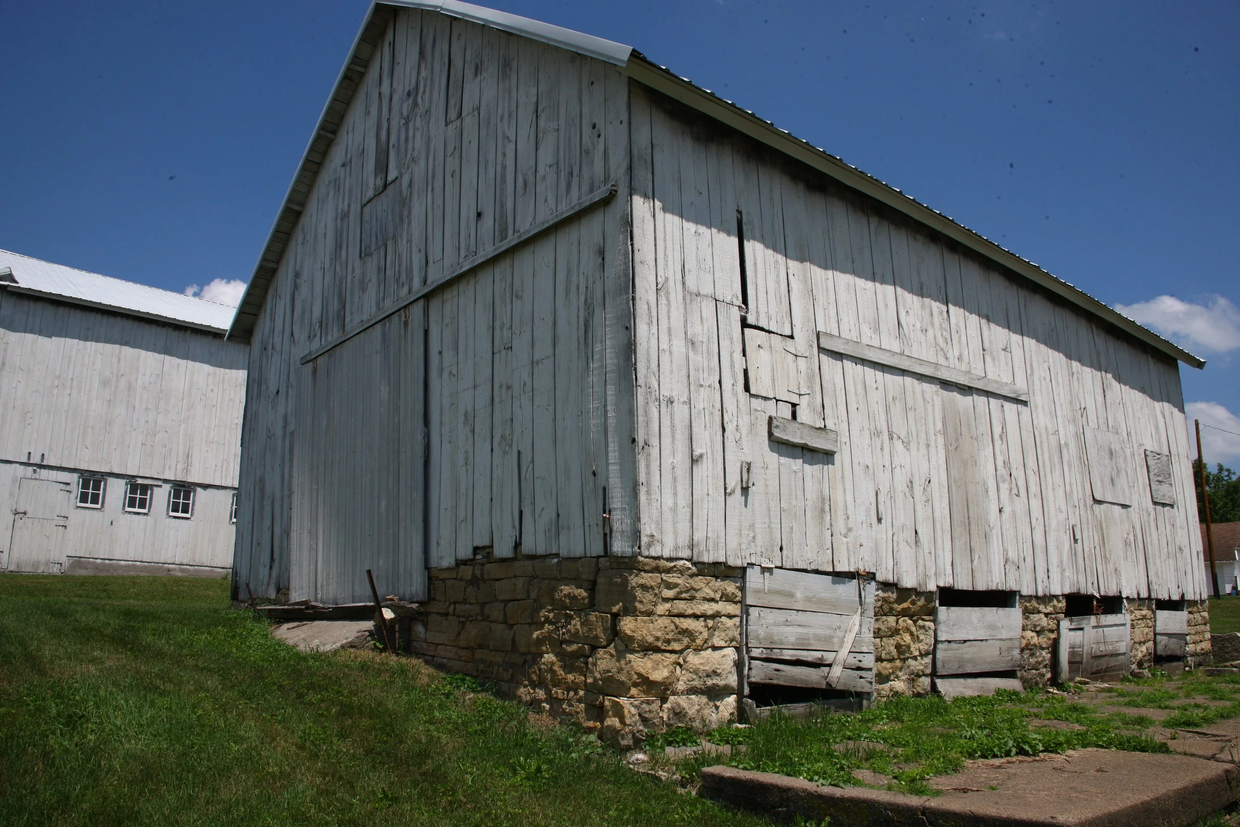 Old white wooden barn with a stone foundation, some boards are broken or missing, under a blue sky.