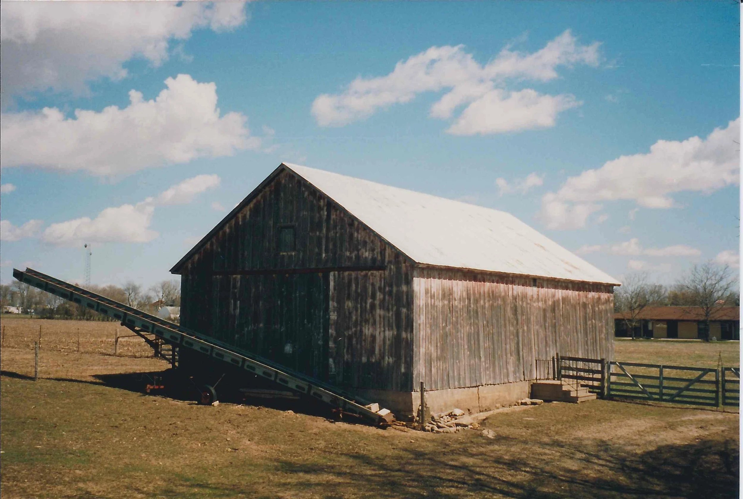 A weathered wooden barn with a white roof and a metal ramp leaning against it, situated on a grassy field under a partly cloudy blue sky.