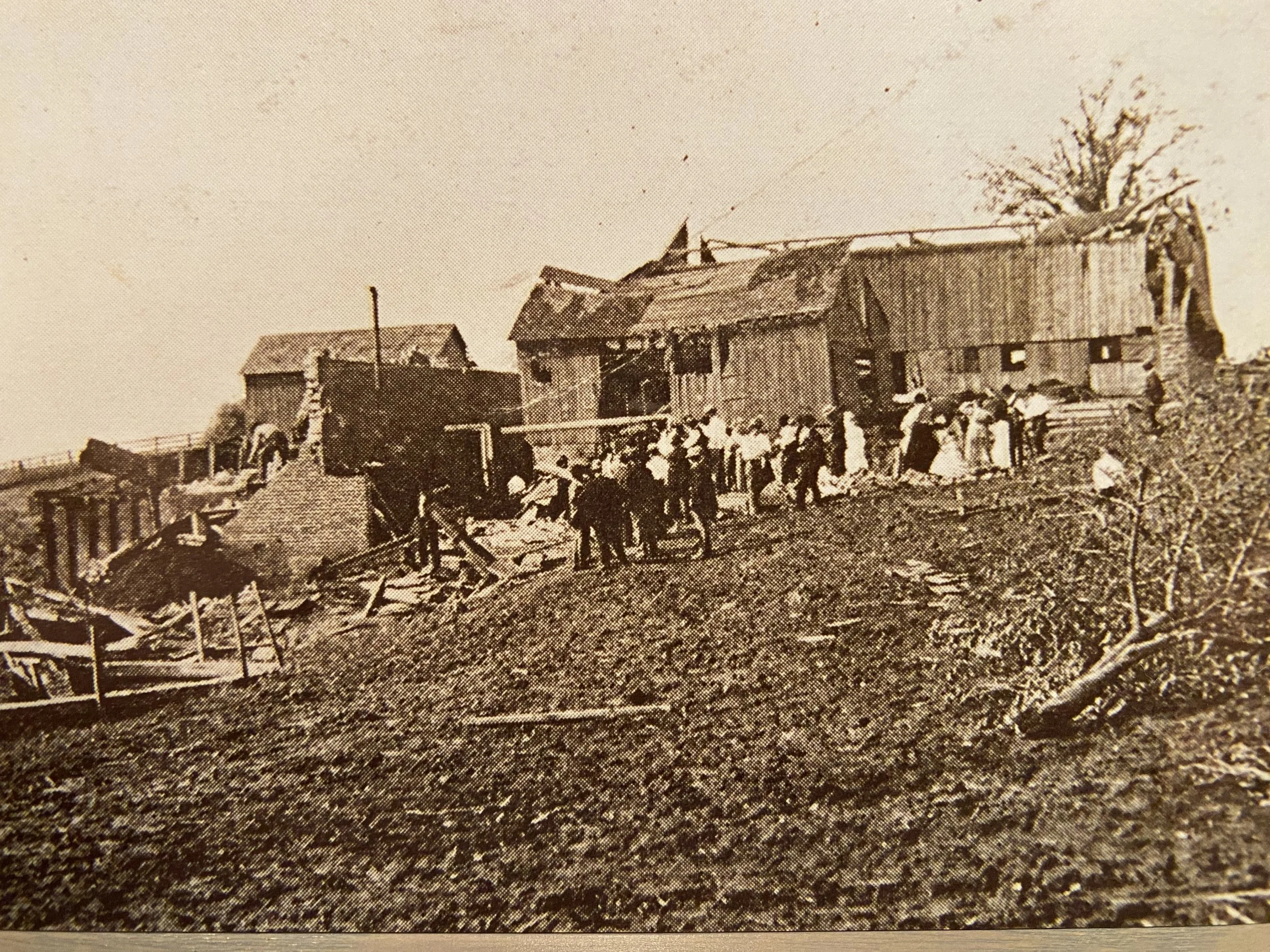 Black and white historical photograph of a damaged wooden building with a group of people gathered outside, some sitting and some standing, on a hilly terrain with debris scattered around.